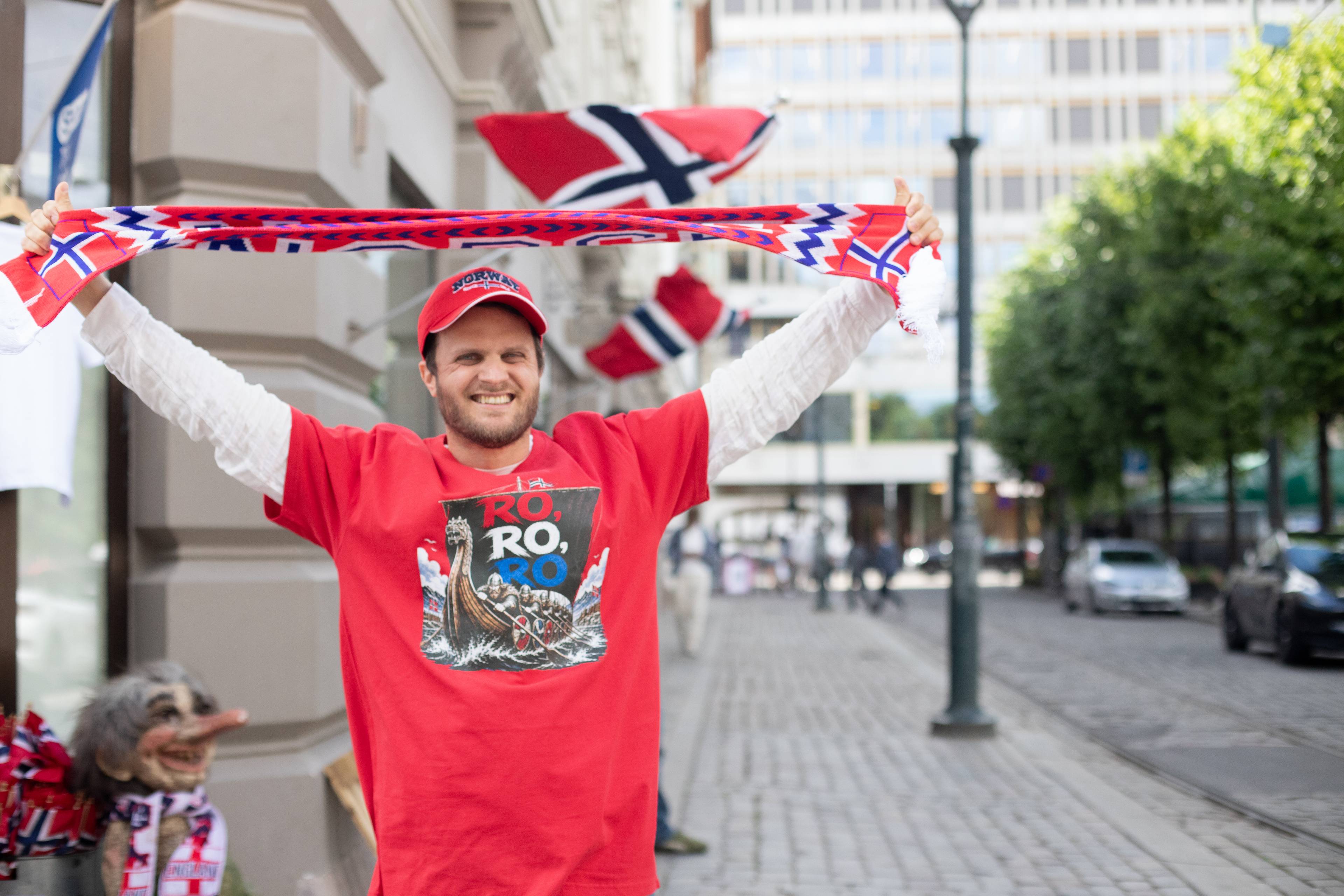 en person som holder et supporter skjerf for Norge, han har på seg en rød t-skjorte der det står "ro, ro, ro" med et motiv av et vikingskip