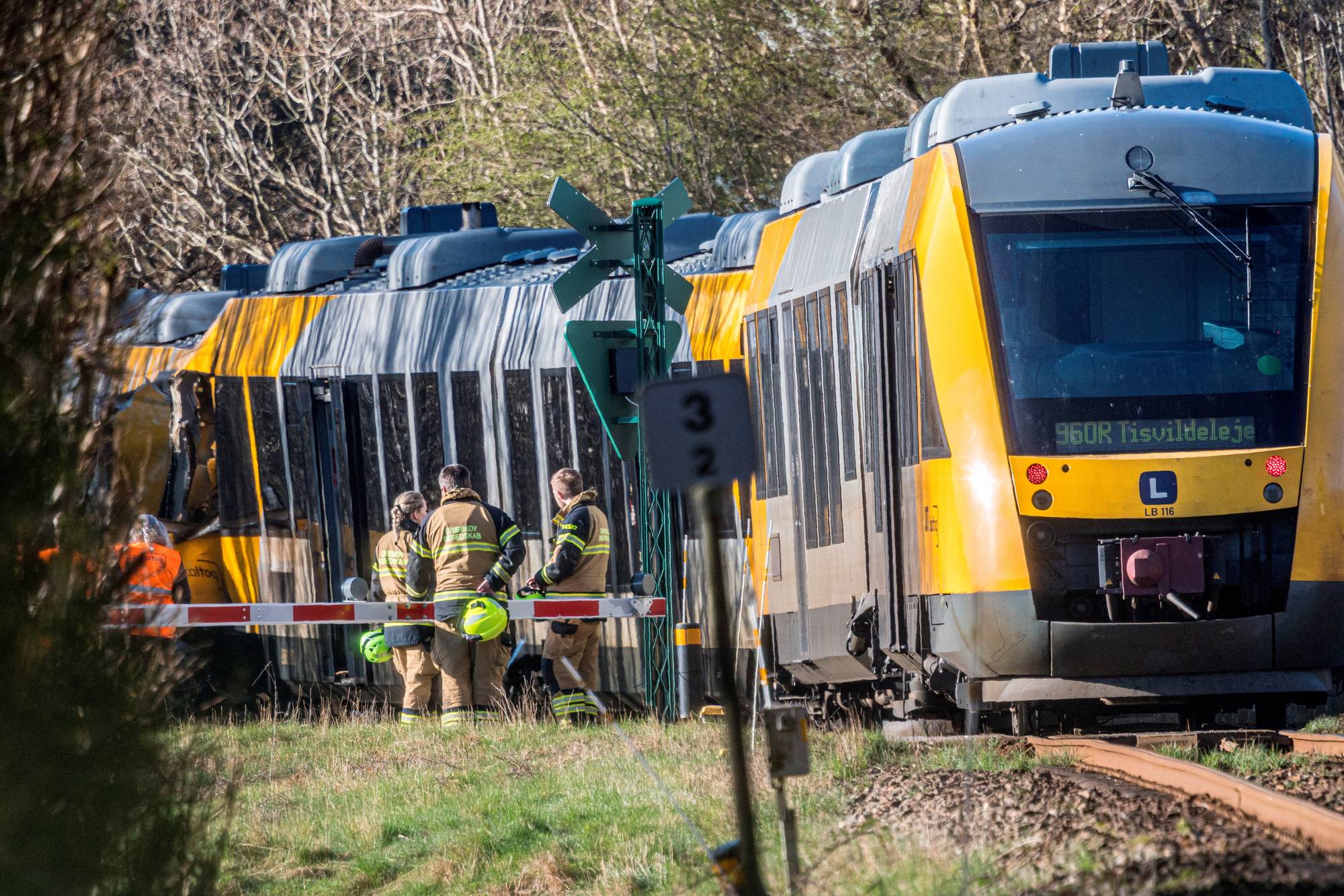 Et tog har sporet av ved en jernbaneovergang. Flere personer i brannmannsutstyr er til stede ved ulykkesstedet. Toget, som er gult og grått, er synlig utenfor sporet. Vegetasjon og trær omkranser området. (Bildebeskrivelsen er laget av en KI-tjeneste)