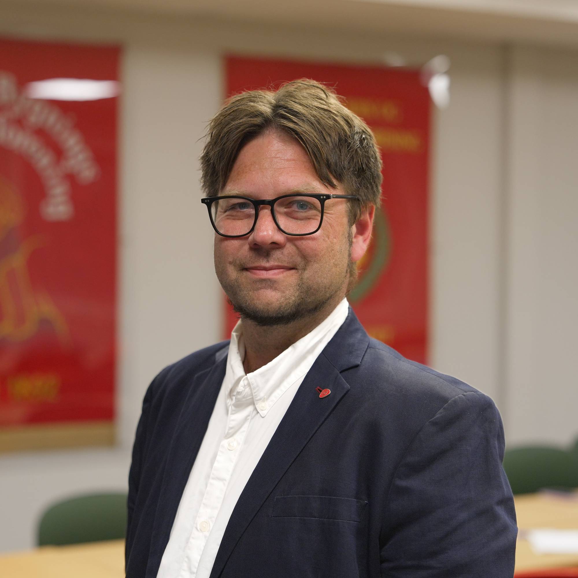 Portrett av mann med briller i mørk jakke, foran røde faner i et møterom. (Bildebeskrivelsen er laget av en KI-tjeneste)