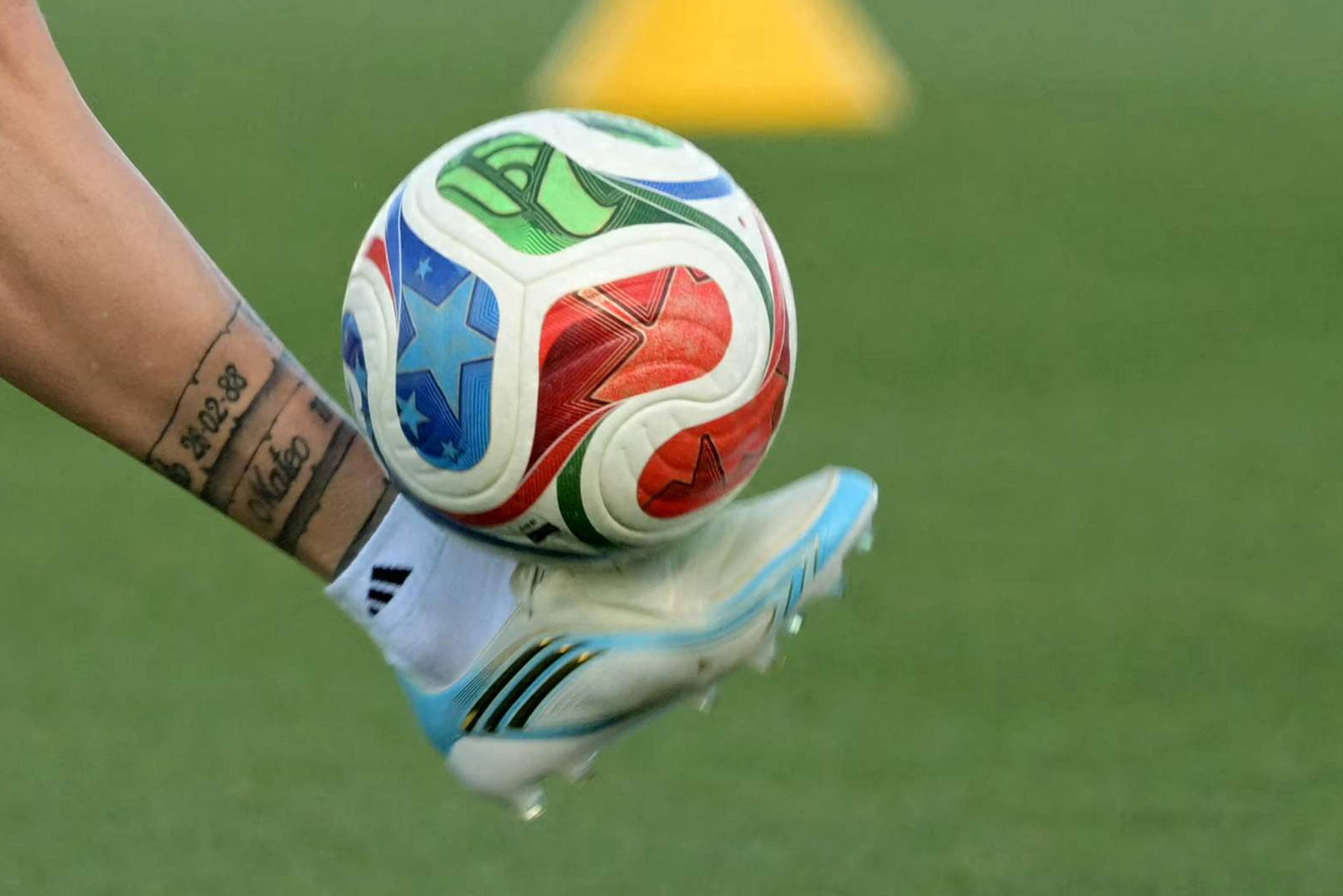 Fotballspiller Lionel Messi trener med ball på en gressbane. Han har på seg hvite og lyseblå drakter og sko. Det er gule kjegler spredt utover banen. I bakgrunnen er det et blått banner med tekst og et hvitt banner med tekst. (Bildebeskrivelsen er laget av en KI-tjeneste)