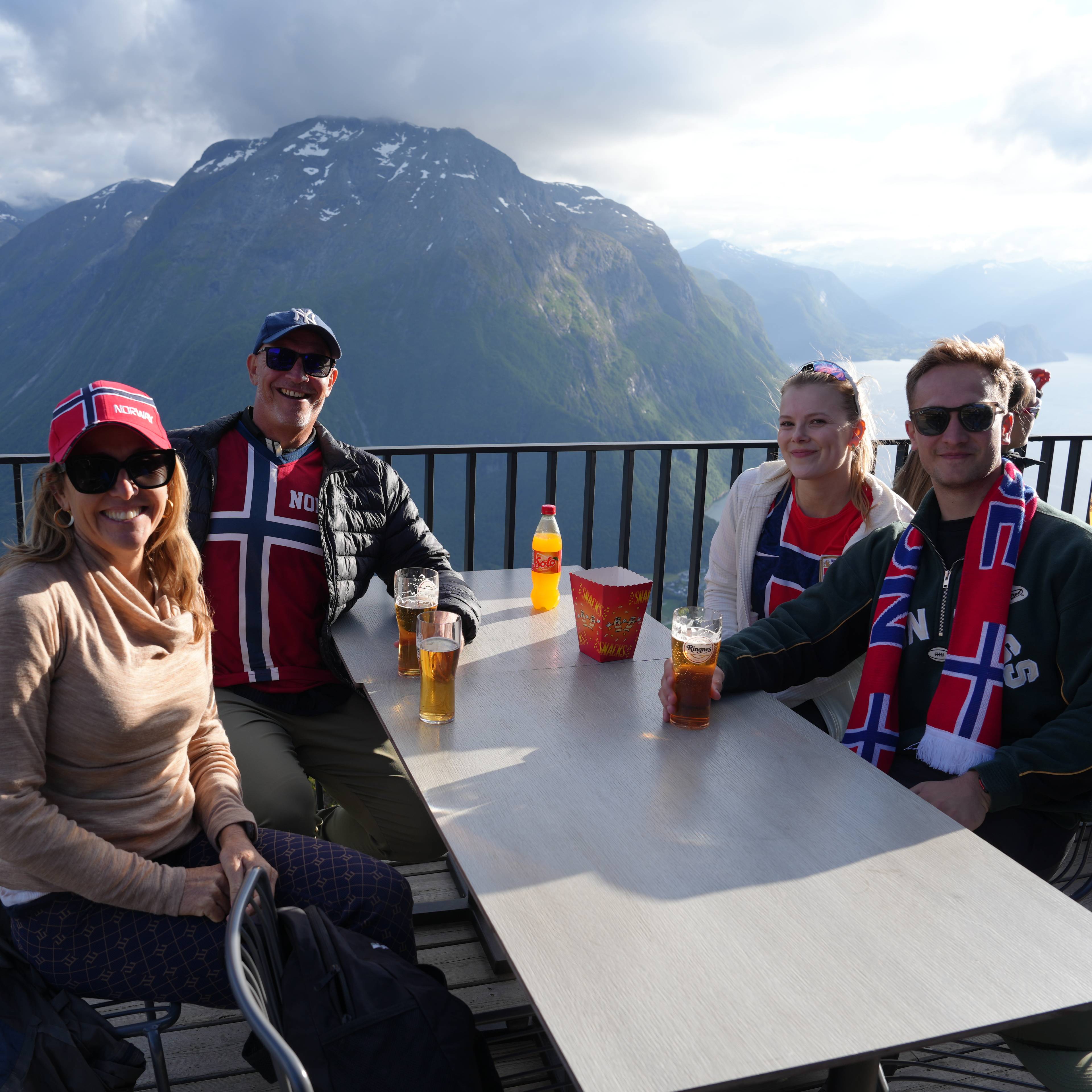 Fire personer sitter ved et bord på en restaurant med fjell og fjord i bakgrunnen. Alle har på seg norsk-inspirerte klær. Mennene har på seg en trøye med det norske flagget. Kvinnene har på seg hodeplagg med det norske flagget. Det er briller med øl og en boks popcorn på bordet. (Bildebeskrivelsen er laget av en KI-tjeneste)