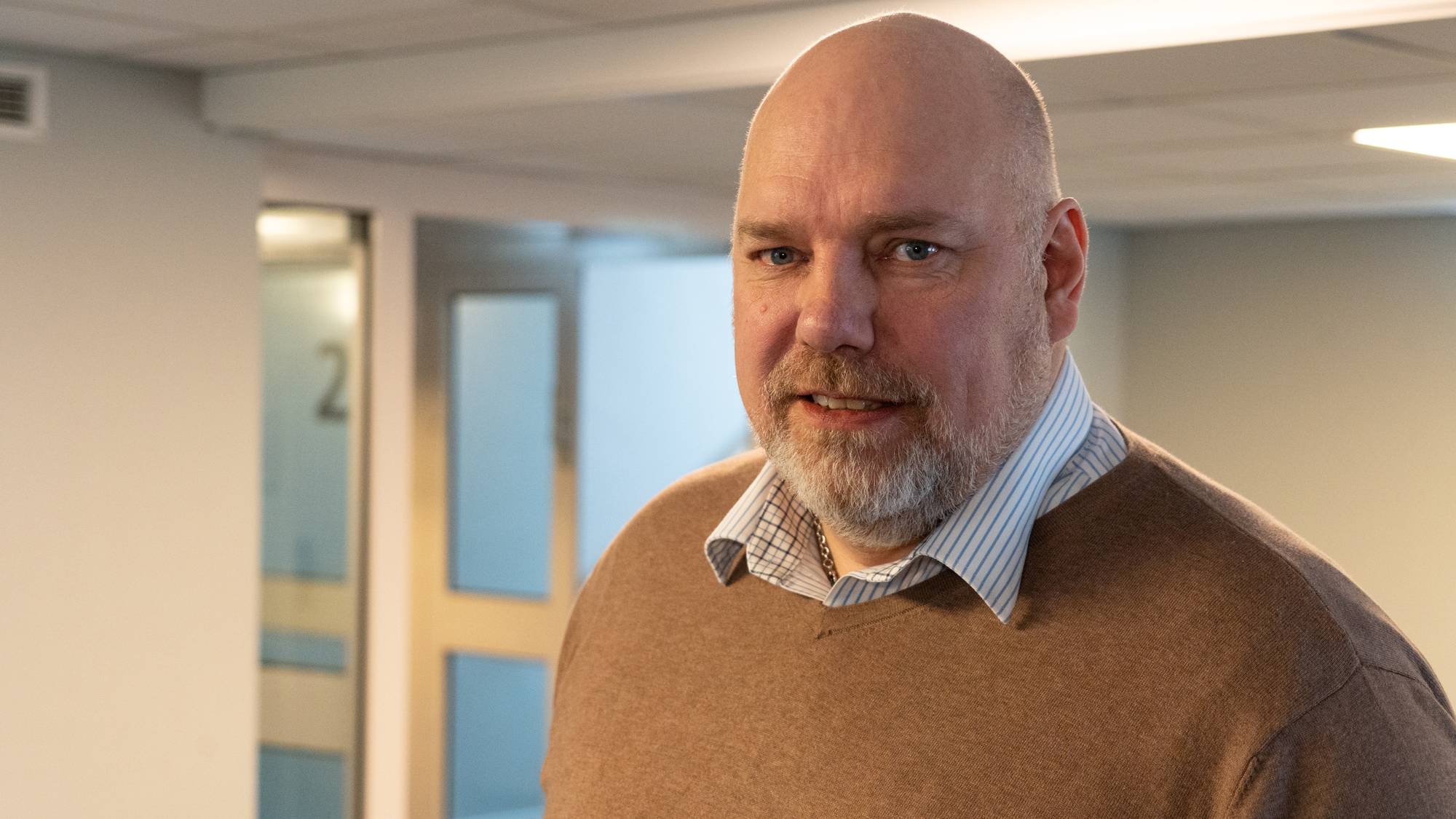 En mann uten hår med brun genser med blå skjorte inni. 