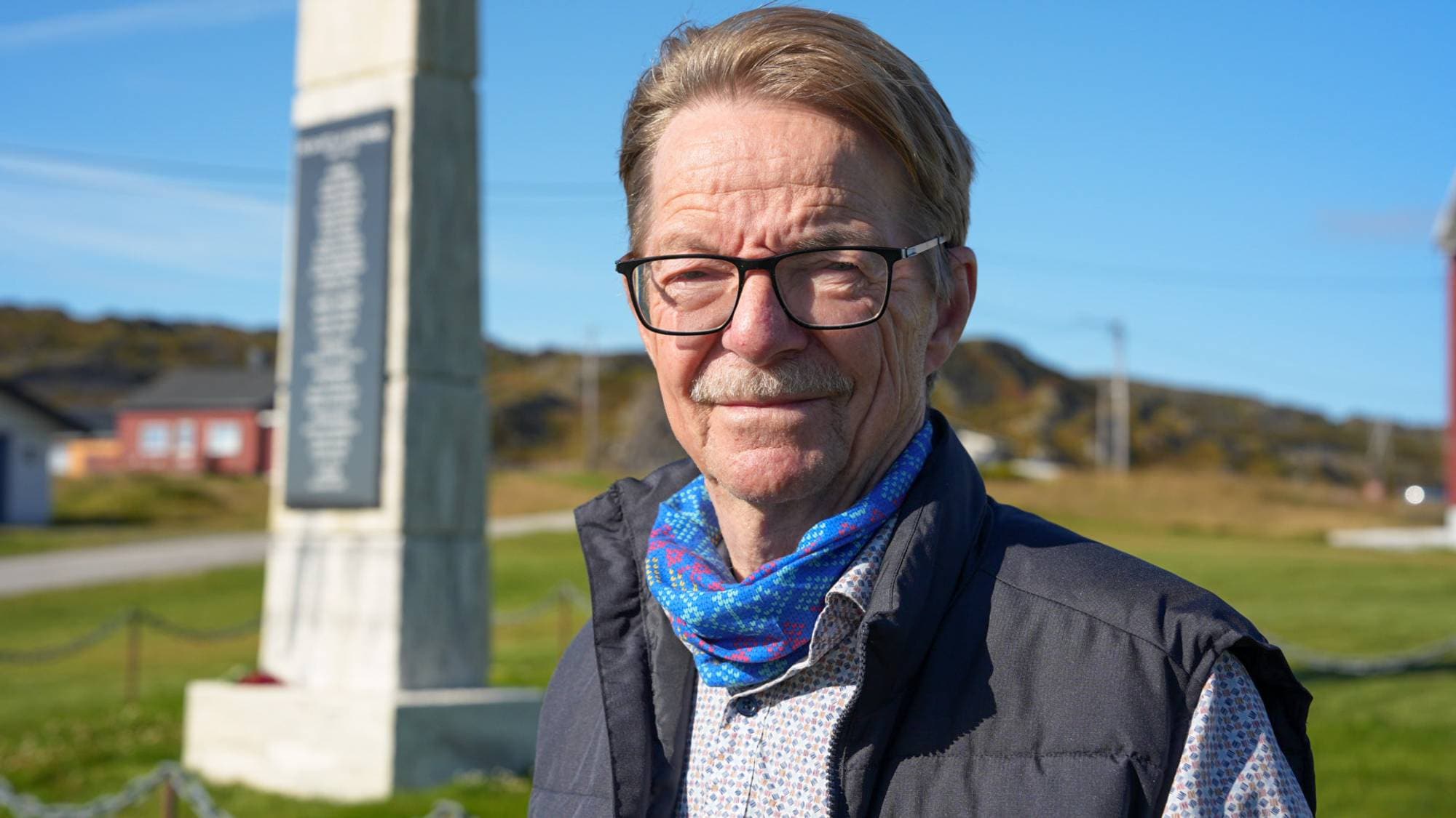 Eldre mann med briller og mønstret skjerf står foran et monument i et utendørs landskap. (Bildebeskrivelsen er laget av en KI-tjeneste)