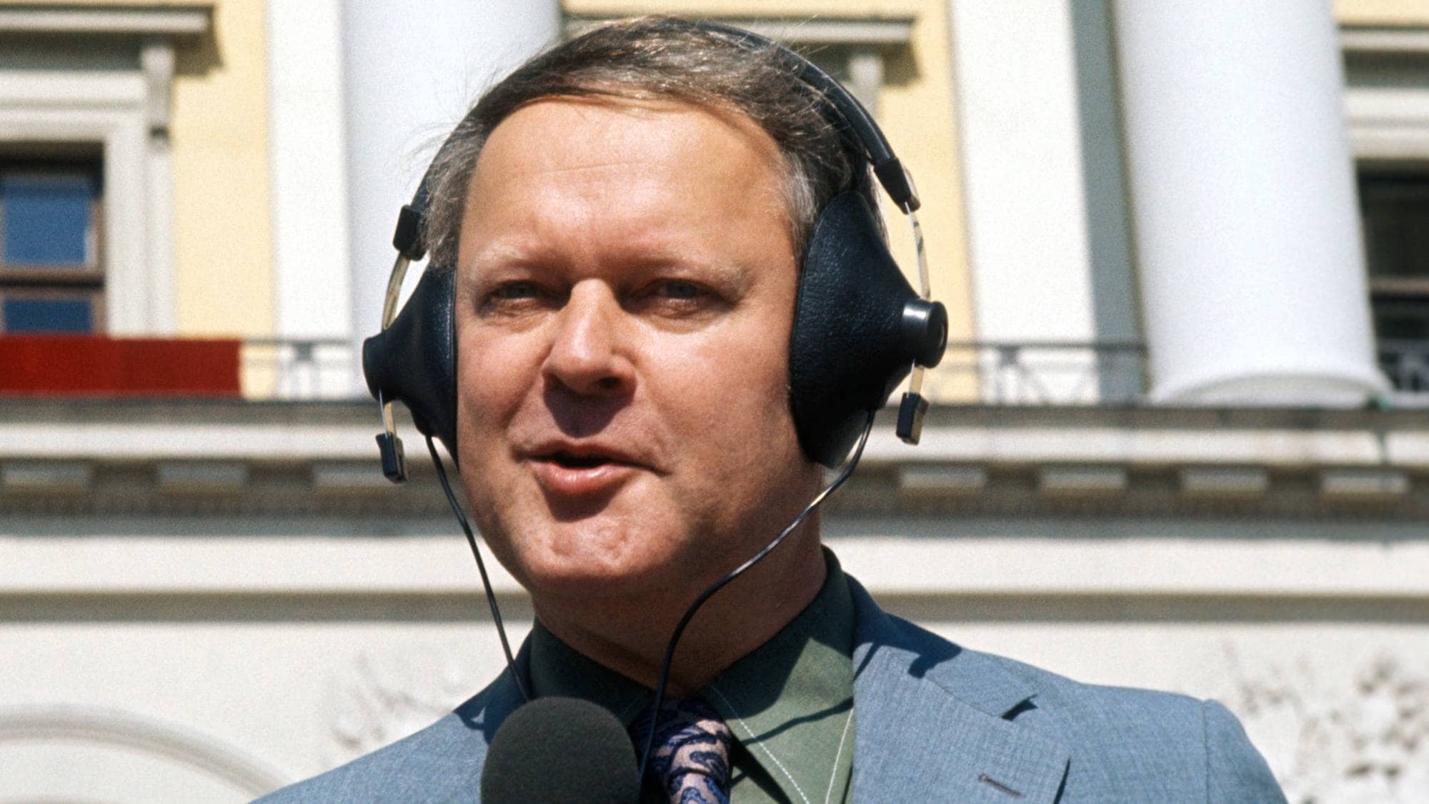 Mann med hodetelefoner og mikrofon foran Det kongelige slott. (Bildebeskrivelsen er laget av en KI-tjeneste)