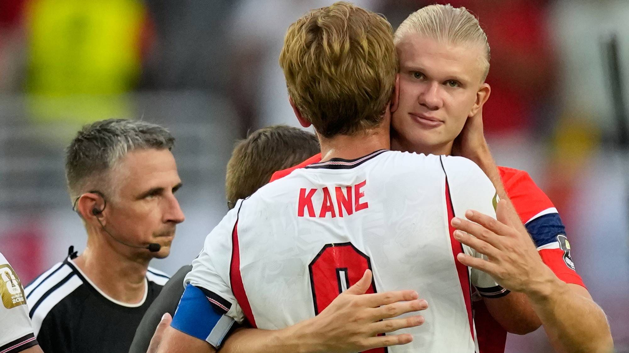 Harry Kane og Erling Braut Haaland klemmer på fotballbanen med dommer og Reece James i nærheten. (Bildebeskrivelsen er laget av en KI-tjeneste)