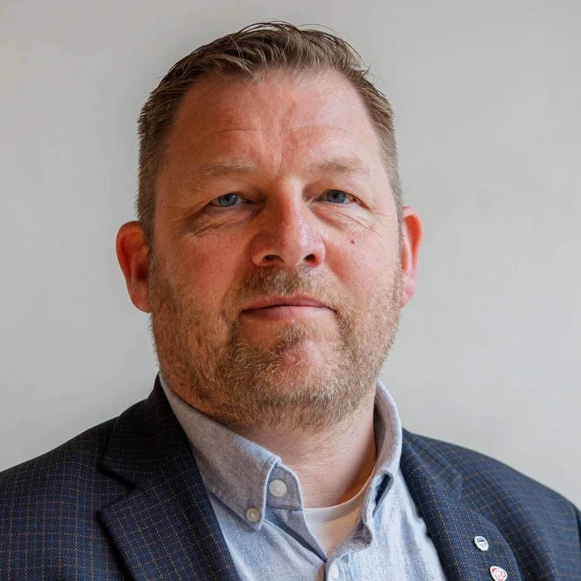 Portrett av en mann med kort hår og skjegg, iført blå skjorte og mørk rutet jakke. (Bildebeskrivelsen er laget av en KI-tjeneste)