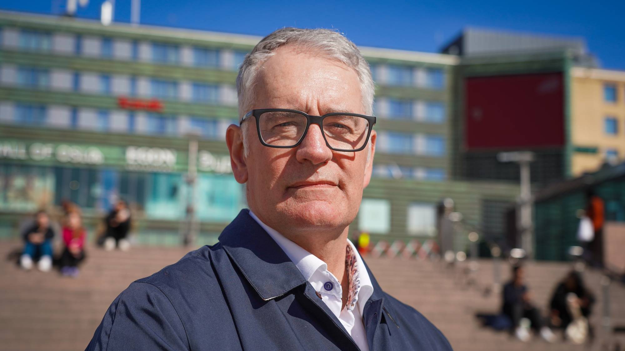 Mann med briller ser mot kameraet. Han er kledd i mørk frakk og hvit skjorte. Bak ham er en bygning med grønn fasade og flere personer som sitter på trapper. Bildet er tatt utendørs i dagslys. (Bildebeskrivelsen er laget av en KI-tjeneste)