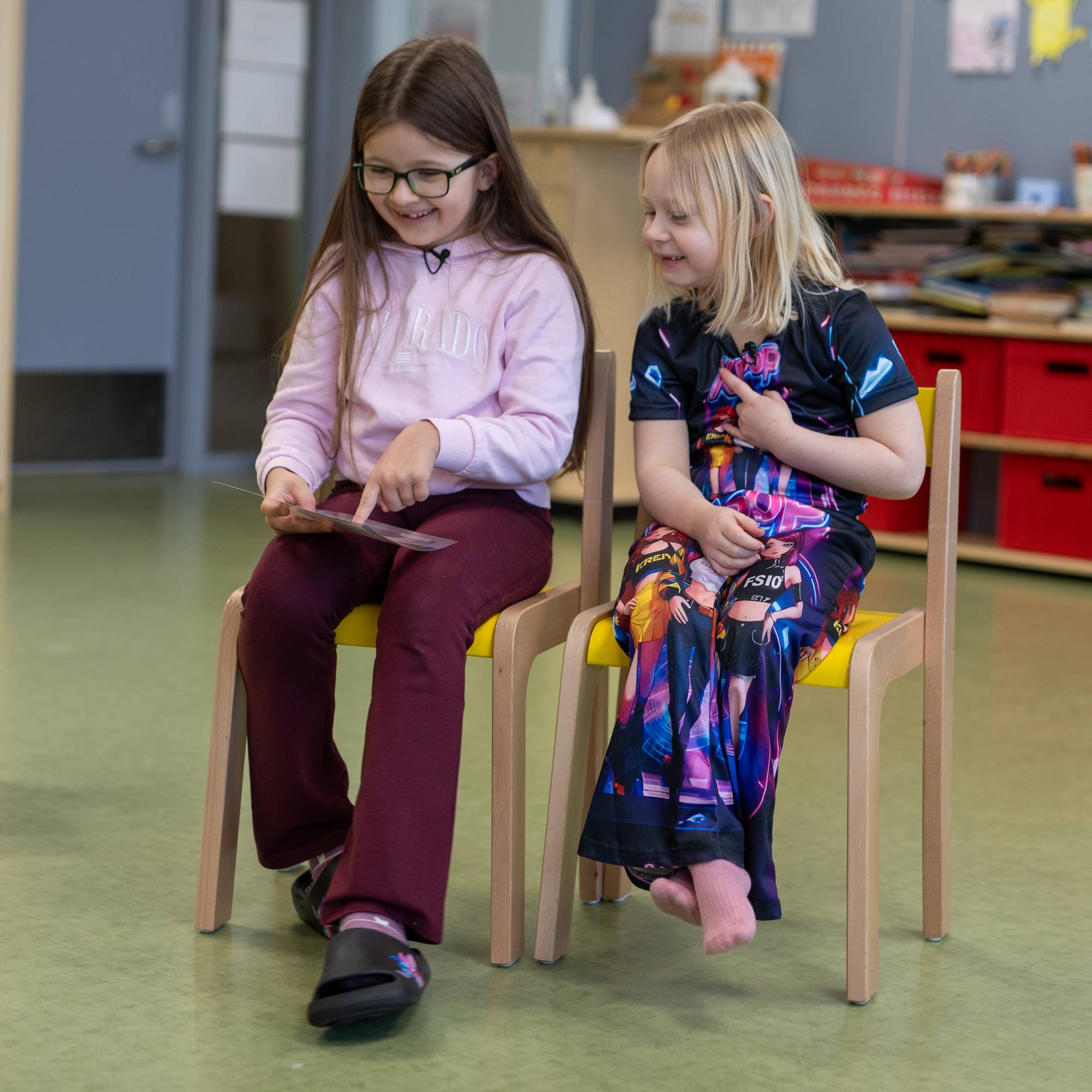 To seks år gamle jenter sitter på stoler inne i et rom på en skole. Den ene peker på et bilde. Begge ser på og smiler.