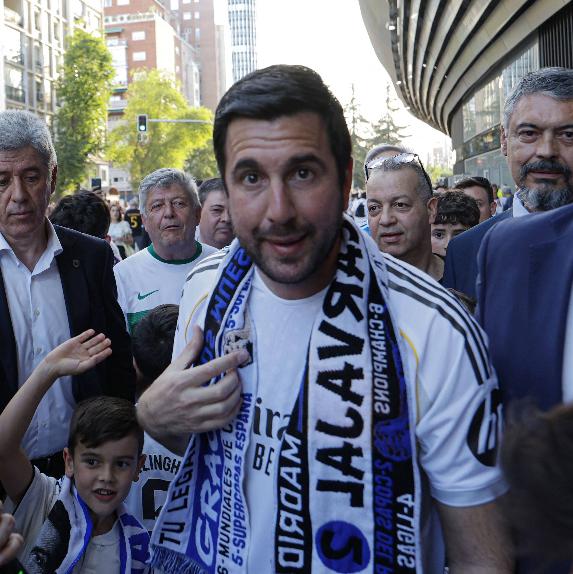 En mann i Real Madrid-drakt med skjerf blir omringet av folkemengde og journalister. En ung gutt vinker til en person med mobiltelefon. Bildet er tatt utendørs med bygninger i bakgrunnen. En person tar bilde av en annen person. (Bildebeskrivelsen er laget av en KI-tjeneste)