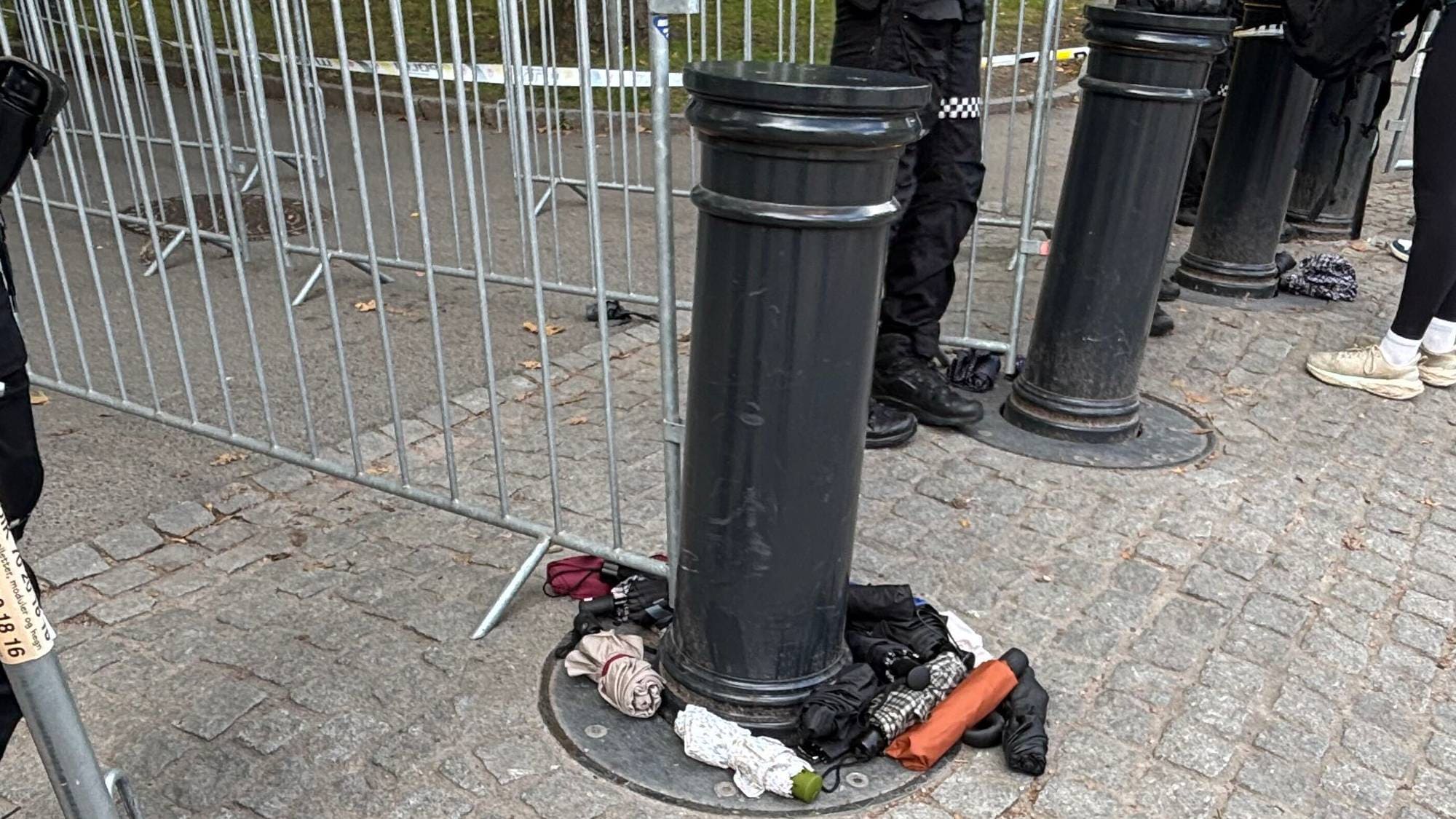 Politi og personer ved metallgjerder utendørs. Paraplyer ligger på bakken ved en stolpe. (Bildebeskrivelsen er laget av en KI-tjeneste)