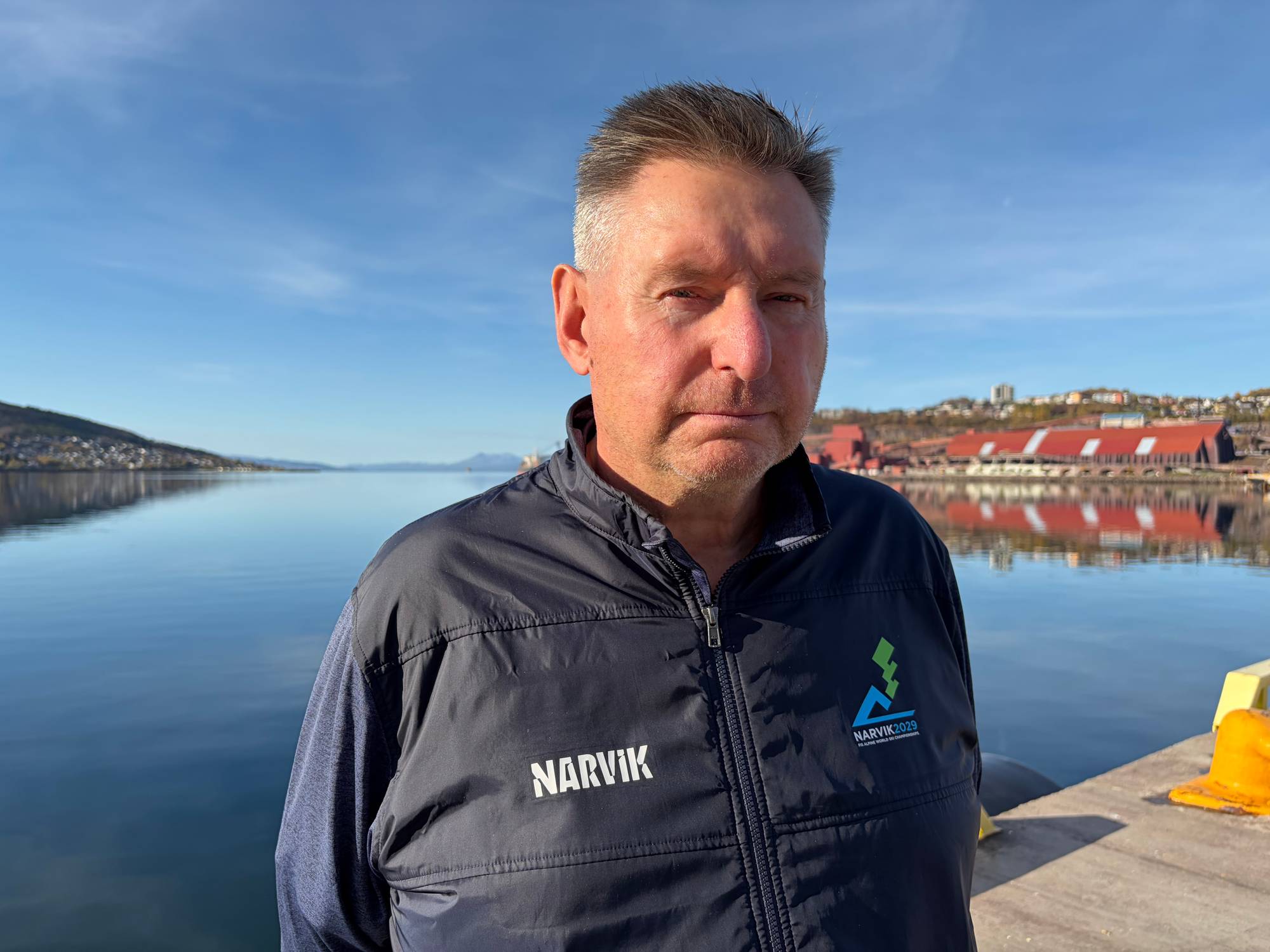 En eldre mann med mørk jakke står ved havna i Narvik med fjell og fjord i bakgrunnen. (Bildebeskrivelsen er laget av en KI-tjeneste)