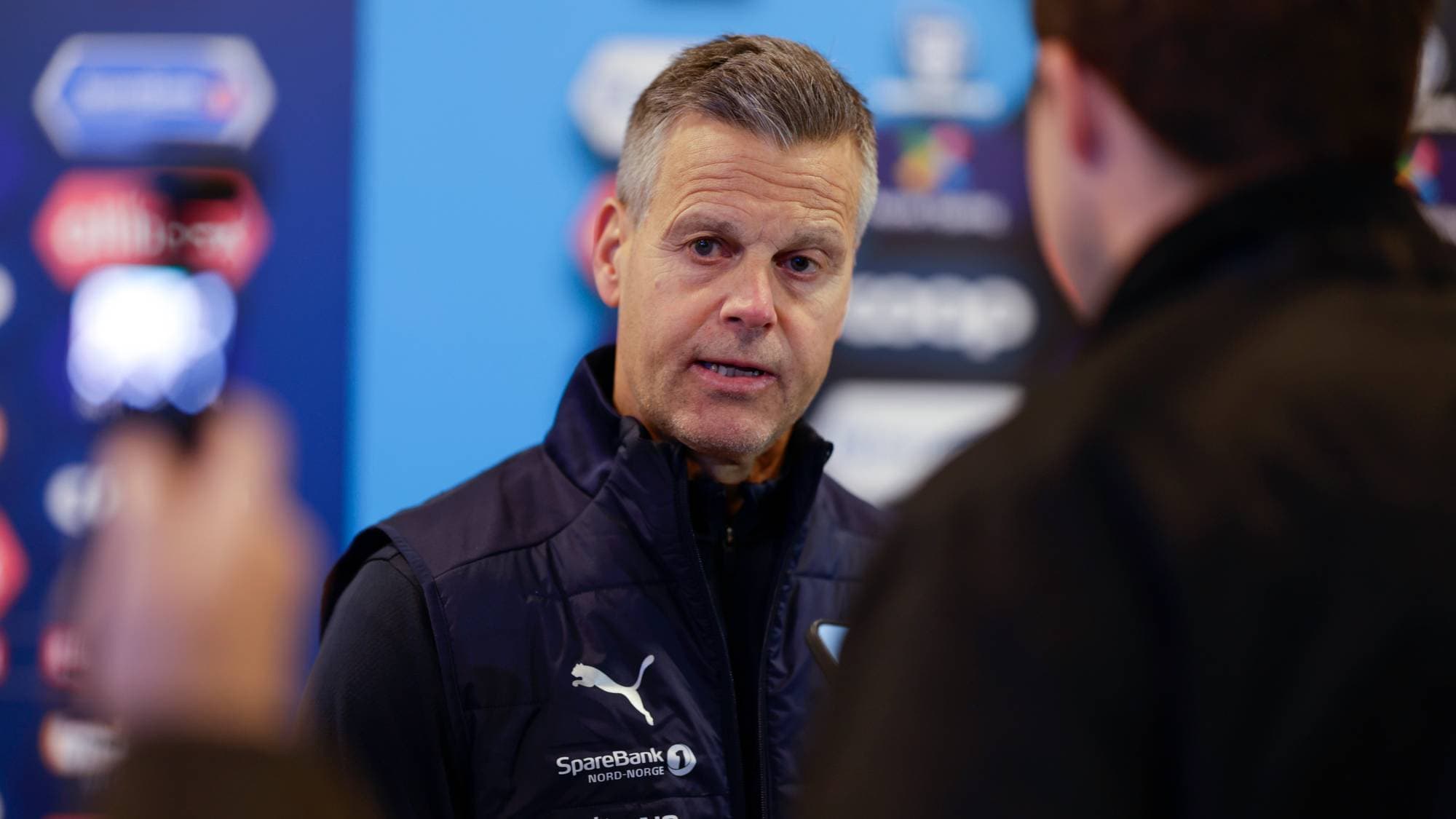 En mann med gråsprengt hår og blå vest blir intervjuet. Han har en Puma-logo og sponsorlogoer på vesten. Bak ham er en blå bakgrunn med forskjellige logoer og tekster. (Bildebeskrivelsen er laget av en KI-tjeneste)