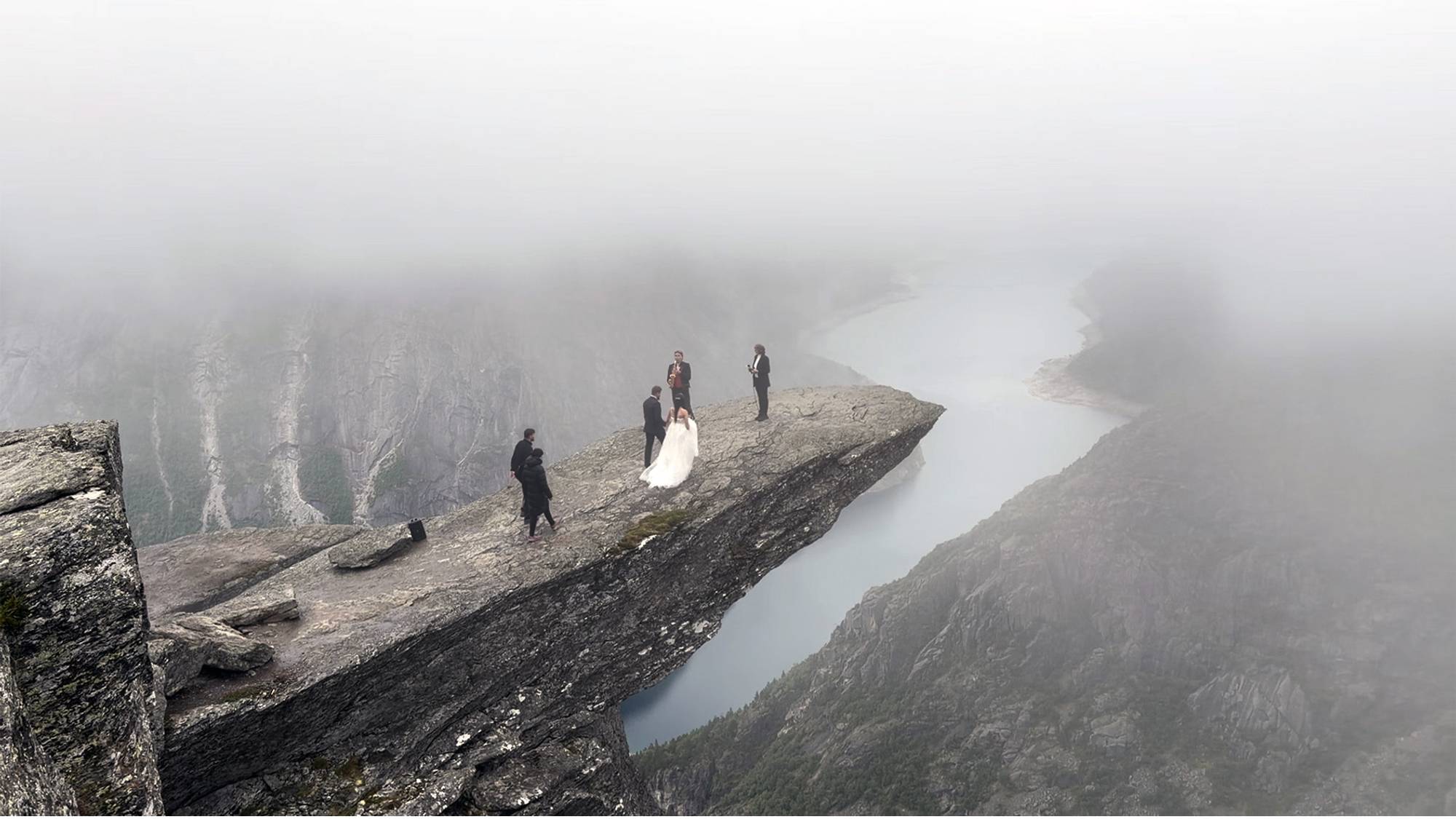 Brurepar på veg ut på Trolltunga for å gifta seg. Forlovarar, og ein musikar er også på steinformasjonen. I bakgrunnen ser ein eit vatn og fjellformasjonar. 