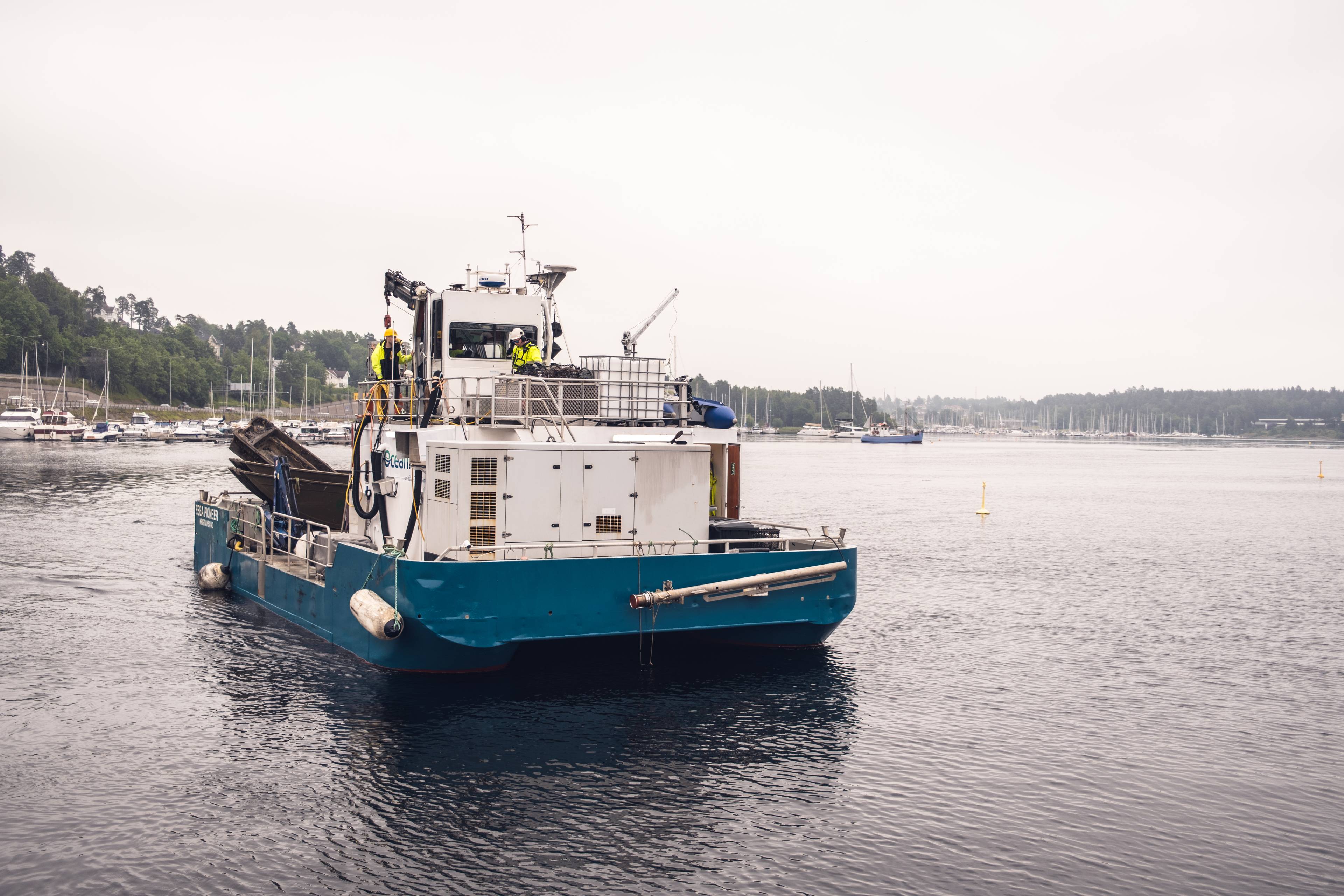 Et blått og hvitt fartøy med mannskap i gule vester manøvrerer i rolig vann. To kraner er synlige på dekk. En gruppe seilbåter og andre fartøy ligger fortøyd i bakgrunnen. En frodig, grønn åsside strekker seg langs vannkanten. (Bildebeskrivelsen er laget av en KI-tjeneste)