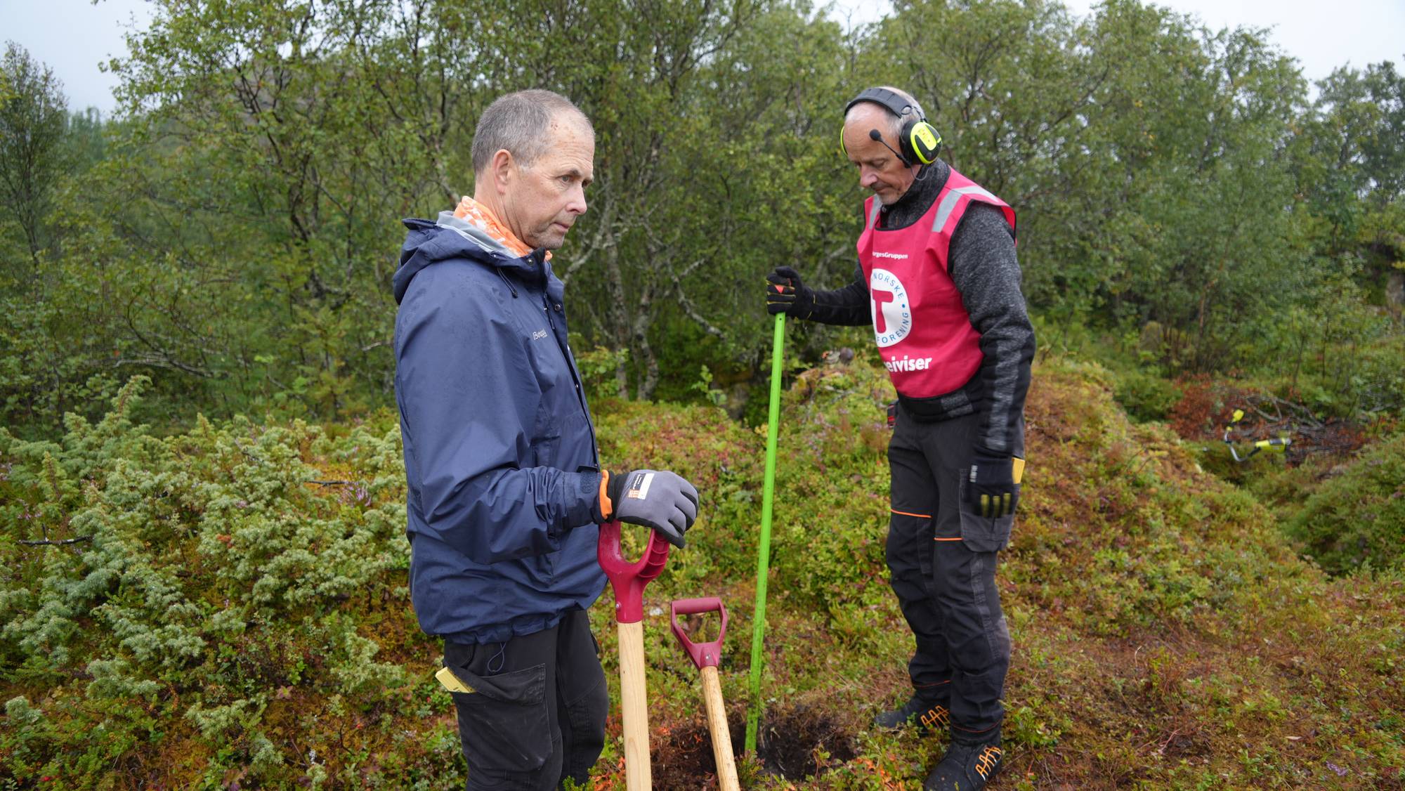 To menn står i et skogterreng med spader, hvor den ene er kledd i vest fra Den Norske Turistforening. (Bildebeskrivelsen er laget av en KI-tjeneste)
