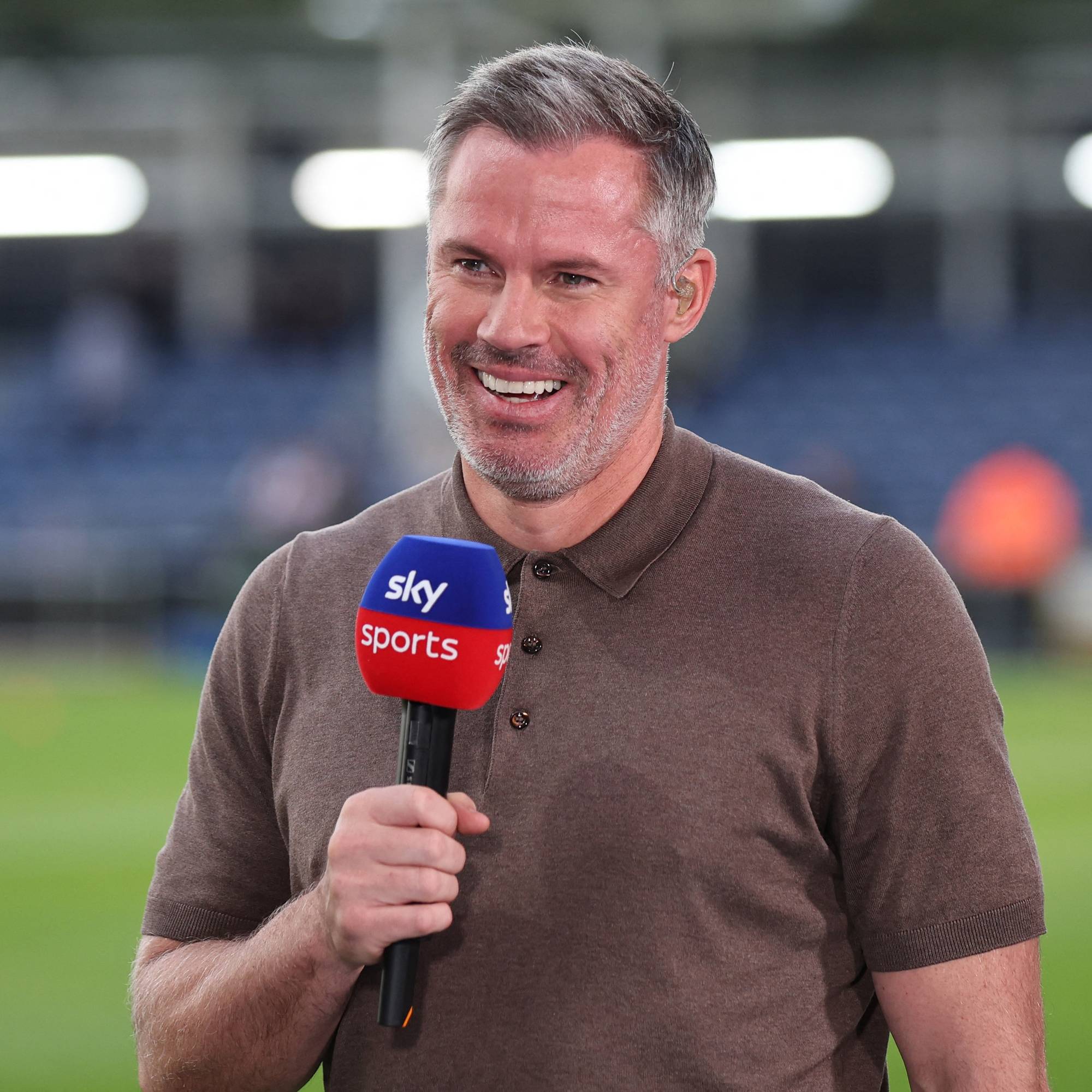 Jamie Carragher holder en Sky Sports-mikrofon mens han smiler foran en fotballbane. (Bildebeskrivelsen er laget av en KI-tjeneste)