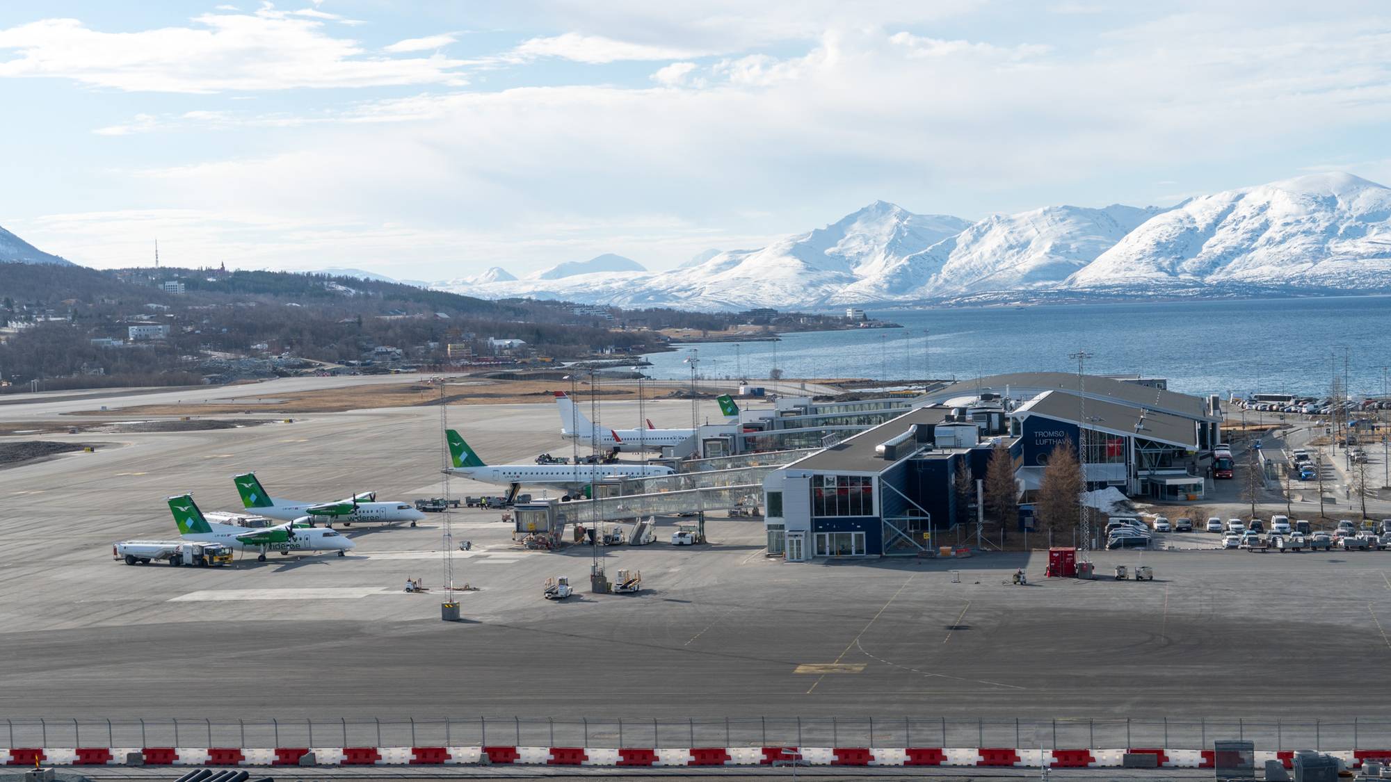 Flyplass med flere fly og en terminalbygning. I bakgrunnen ligger et snødekt fjellandskap ved et islagt hav. Flere biler er parkert ved terminalen. (Bildebeskrivelsen er laget av en KI-tjeneste)
