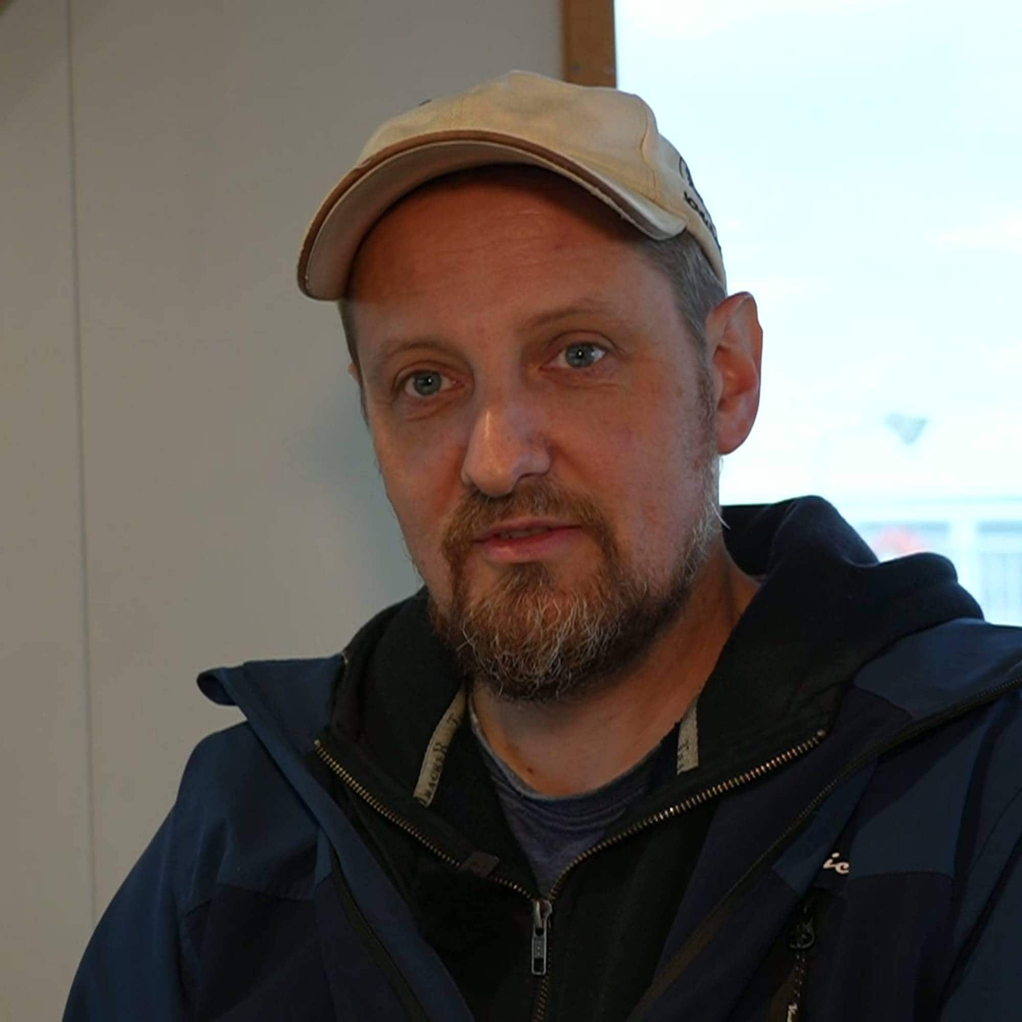 En mann med skjegg og lys caps sitter innendørs i en mørk jakke. (Bildebeskrivelsen er laget av en KI-tjeneste)