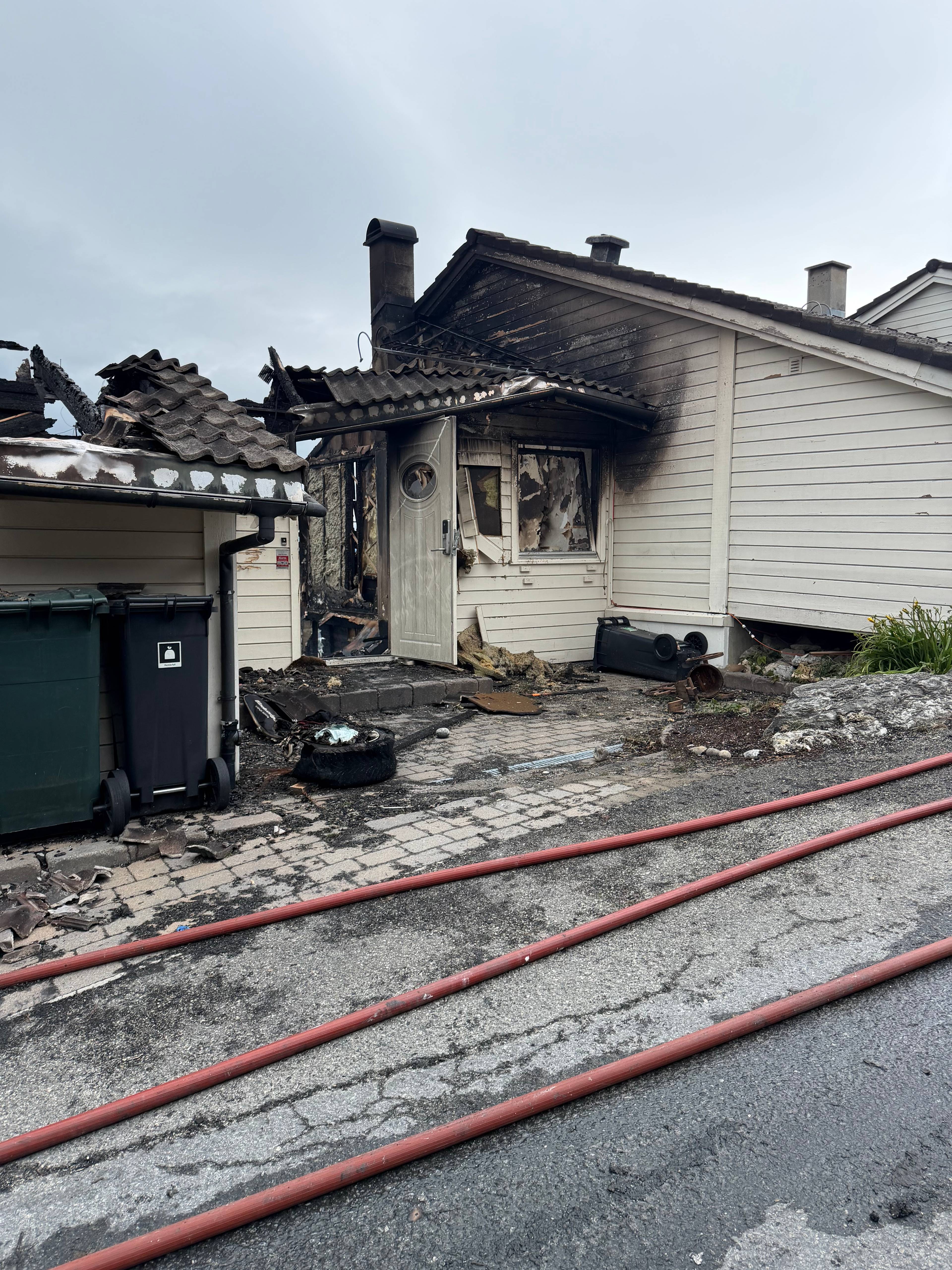 Brannskadet bolig med brente vegger og tak. Røde brannslanger ligger på bakken foran huset. (Bildebeskrivelsen er laget av en KI-tjeneste)