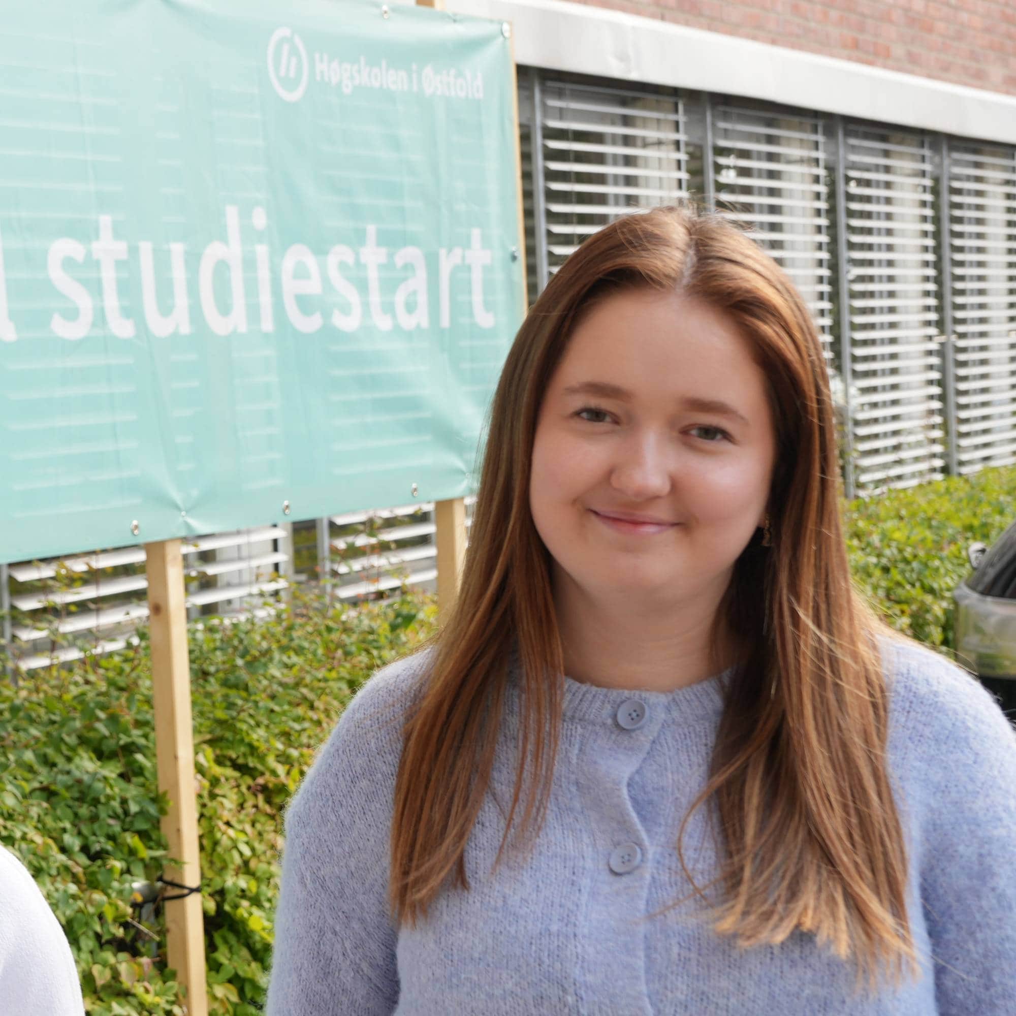 En jente og gutt står utenfor et skilt hvor det står "Velkommen til studiestart". Hun har på seg en blå jakke, og han har på seg en hvit t-skjorte. 