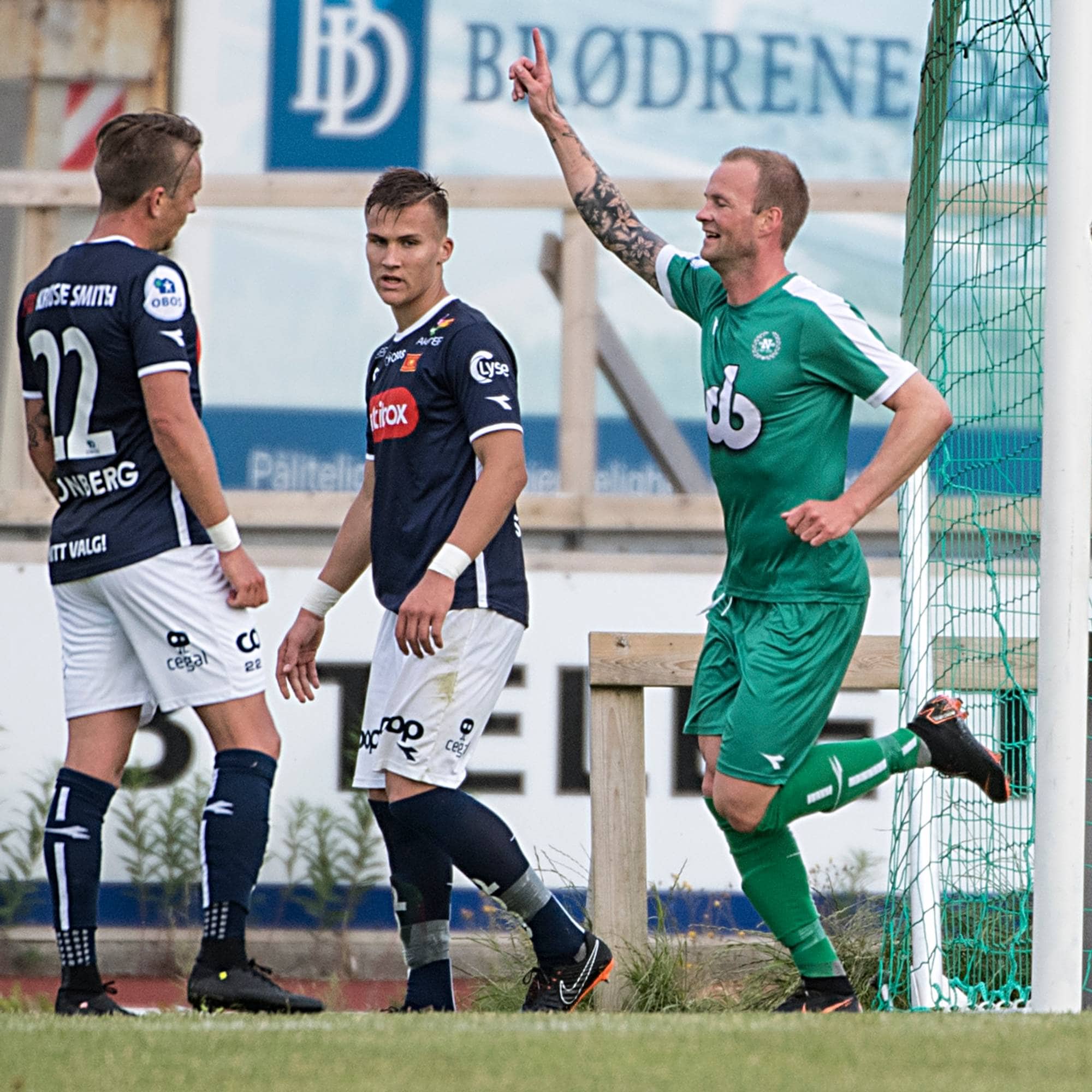 Fotballspiller i grønn drakt feirer mål sammen med medspiller foran motstandere i mørke drakter. (Bildebeskrivelsen er laget av en KI-tjeneste)