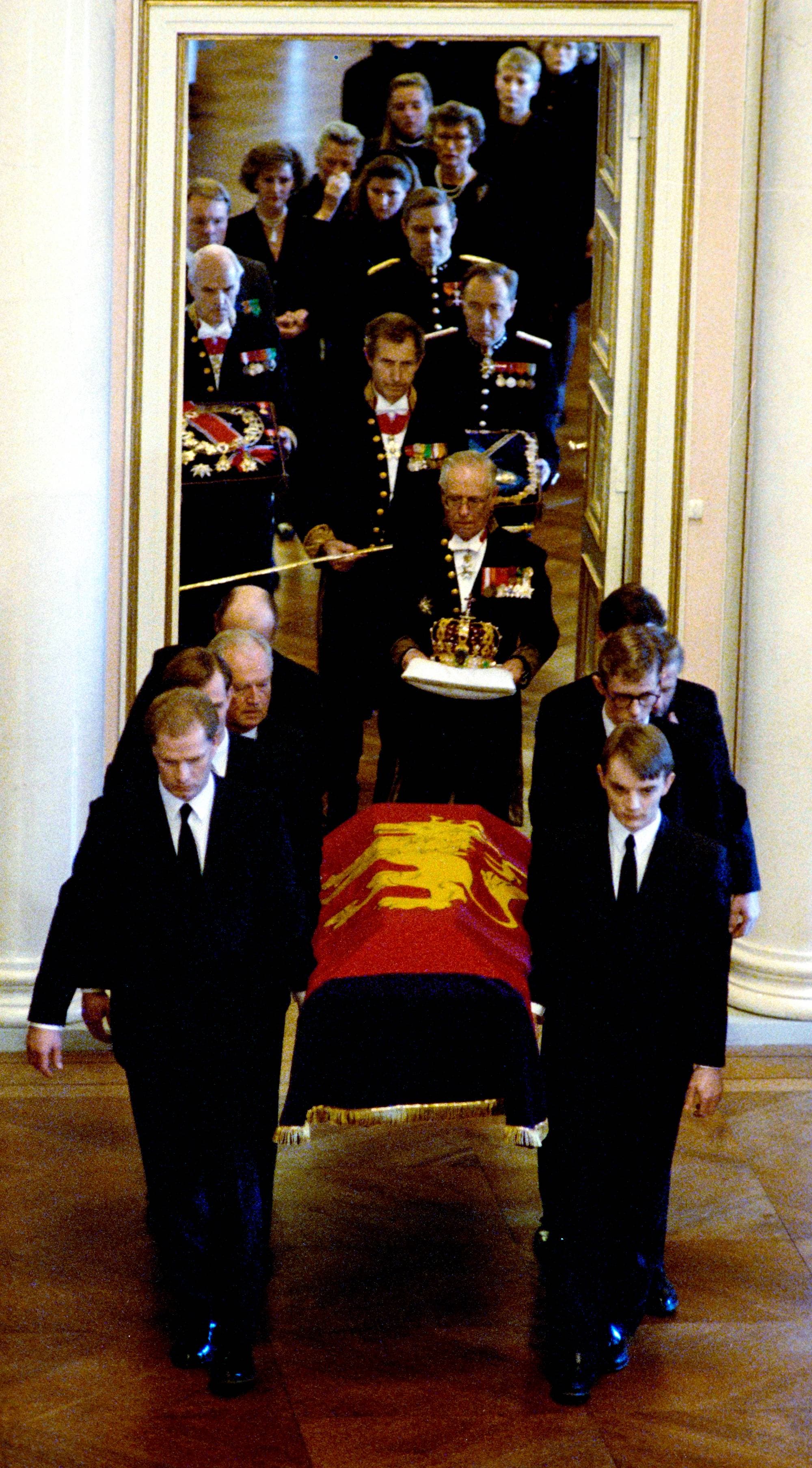 Menn i mørke dresser bærer en kiste dekket av et kongeflagg i en prosesjon i et slott. 