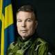 Hampus Andersson / Försvarsmakten En mann i militær uniform ser rett inn i kamera. Det er en mørk bakgrunn og et svensk flagg til venstre bak mannens høyre skulder.