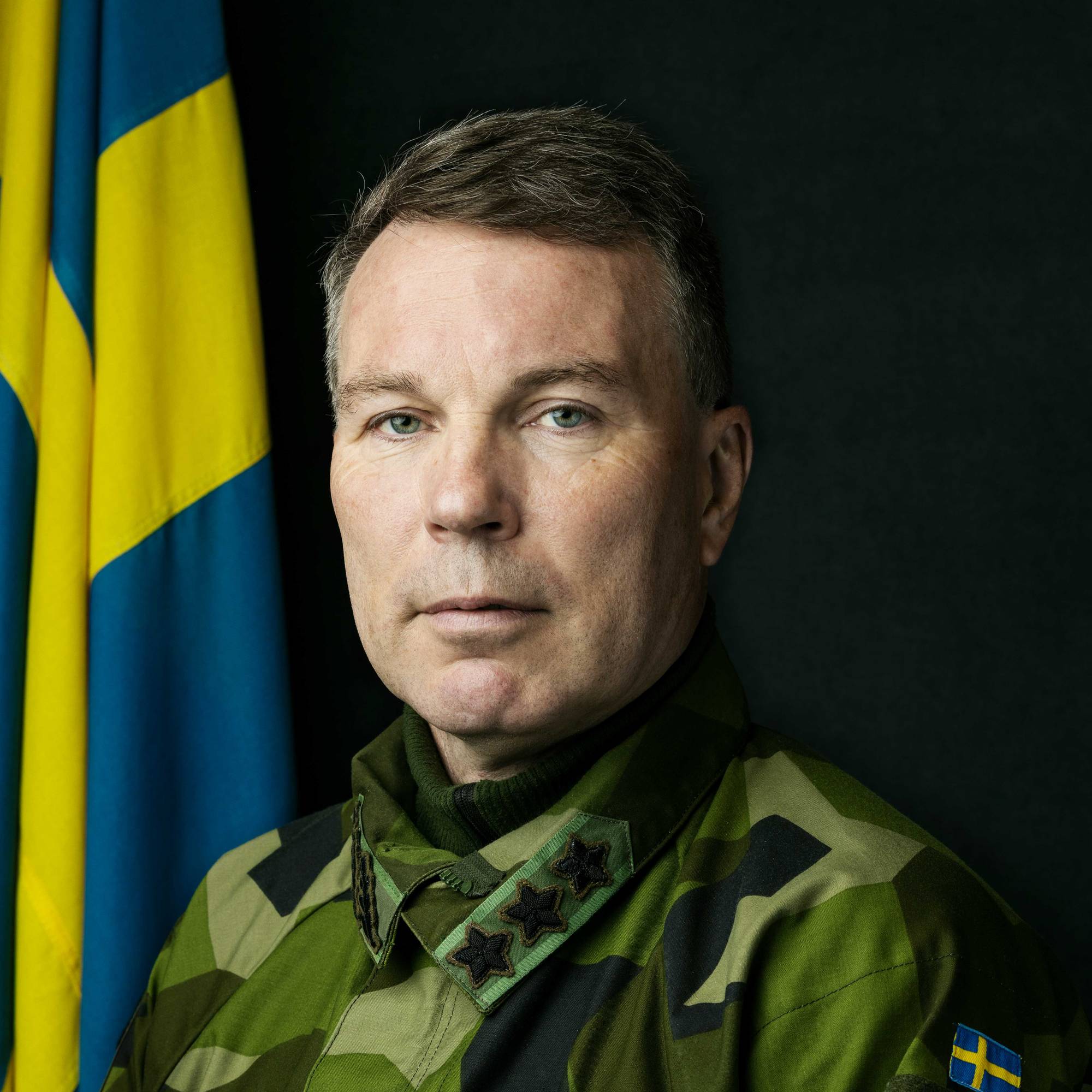 En mann i militær uniform ser rett inn i kamera. Det er en mørk bakgrunn og et svensk flagg til venstre bak mannens høyre skulder.