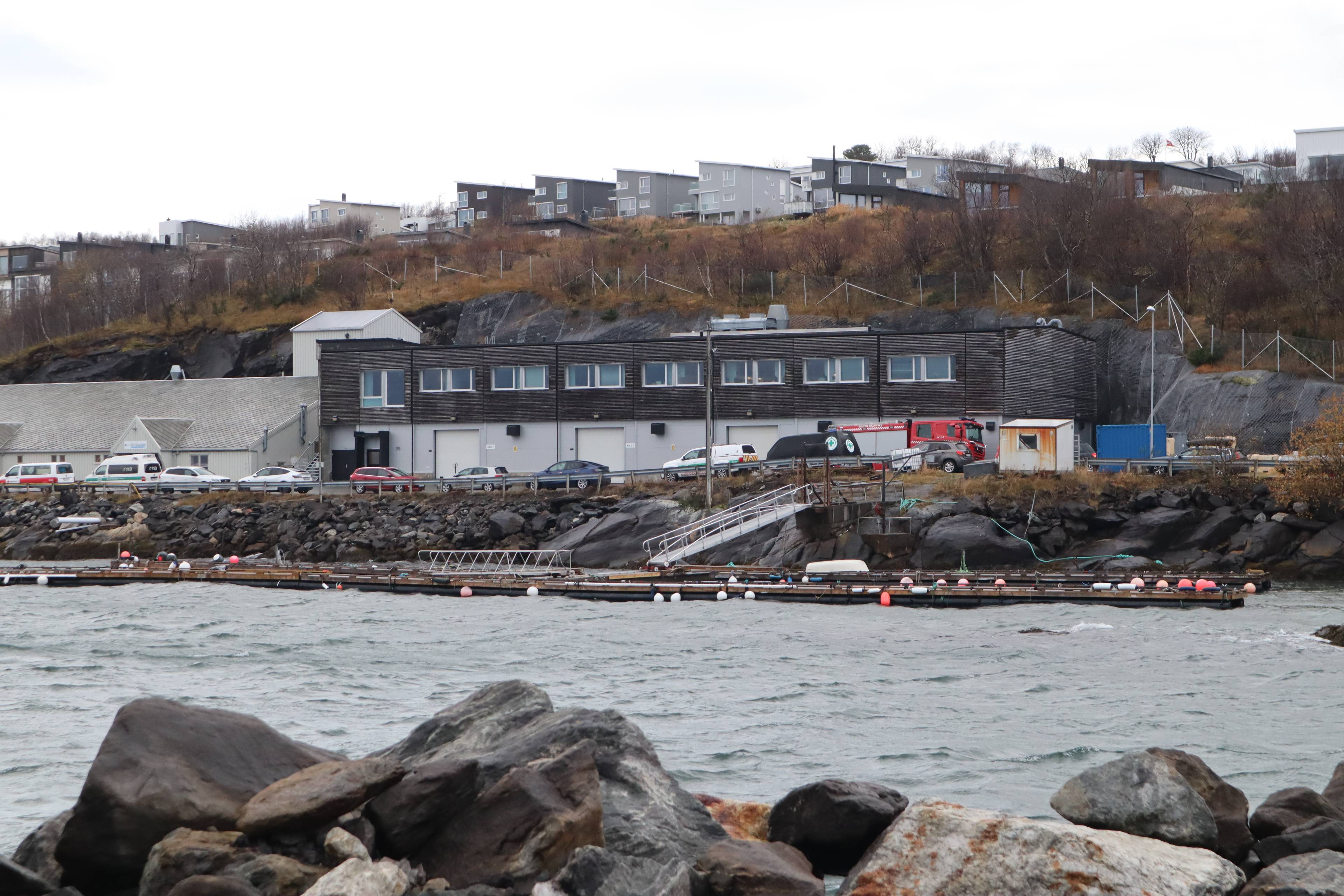 Brannbil og utrykningskjøretøy parkert ved en næringsbygning i en skråning ved sjøen. (Bildebeskrivelsen er laget av en KI-tjeneste)