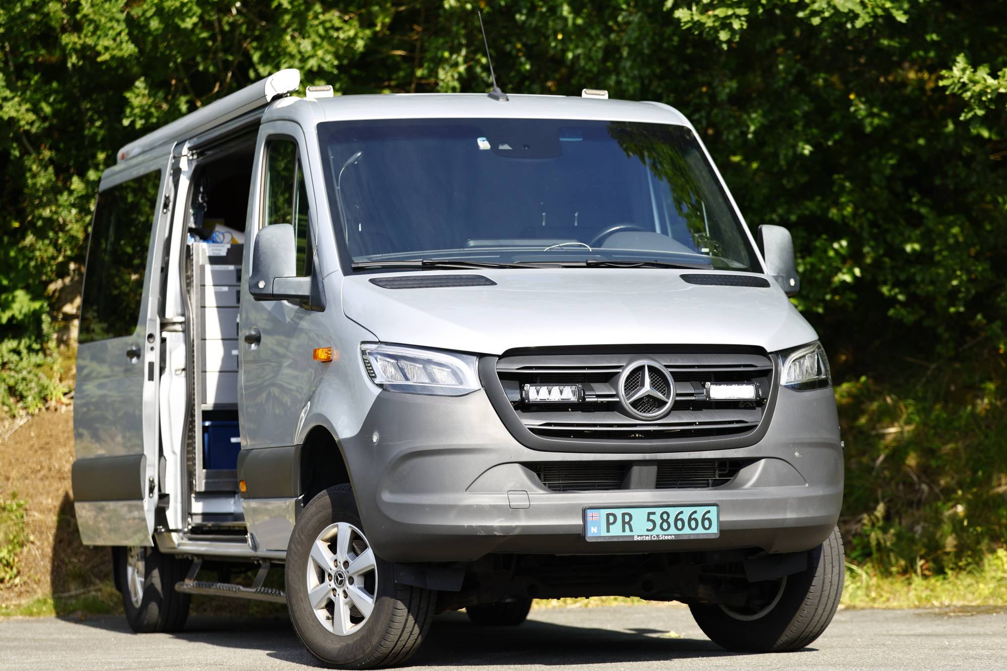 En sølvfarget Mercedes-Benz varebil med åpen skyvedør parkert foran trær. (Bildebeskrivelsen er laget av en KI-tjeneste)