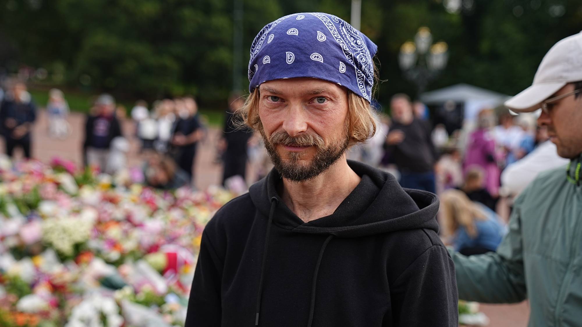 Mann med blått hodeplagg foran blomsterhav ved en minnemarkering. (Bildebeskrivelsen er laget av en KI-tjeneste)