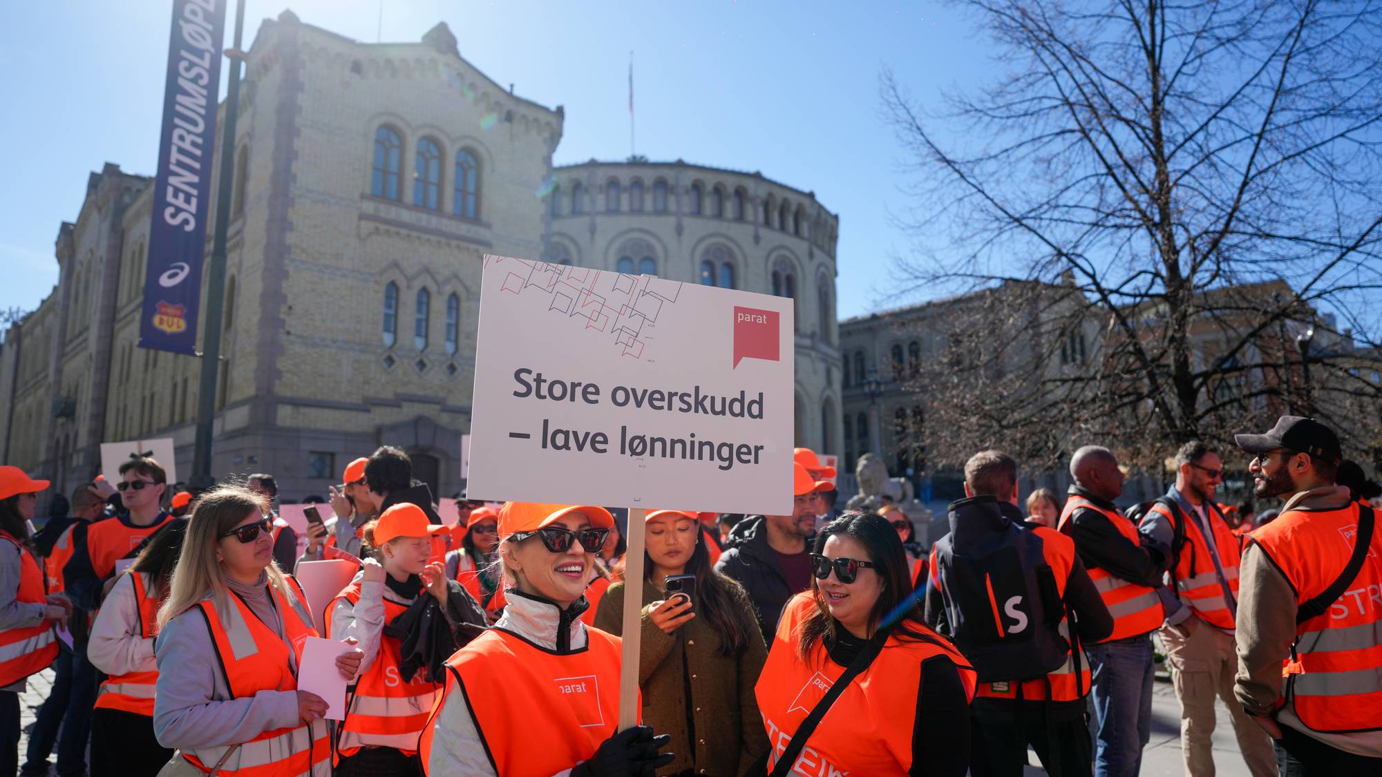 Demonstranter med oransje vester står utenfor Stortinget. De holder skilt der det står "Store overskudd - lave lønninger". Solen skinner og himmelen er blå. En av demonstrantene holder en mobiltelefon og tar bilde. (Bildebeskrivelsen er laget av en KI-tjeneste)