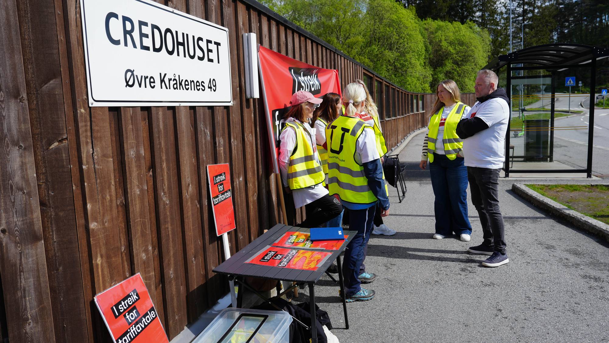 En gruppe personer står ved et skilt med teksten "Credohuset Øvre Kråkenes 49". De er iført gule vester og flere holder opp skilt hvor det står "I streik for tariffavtale". Bak dem henger et rødt banner med lignende budskap. I bakgrunnen ses en bussholdeplass og trær. 