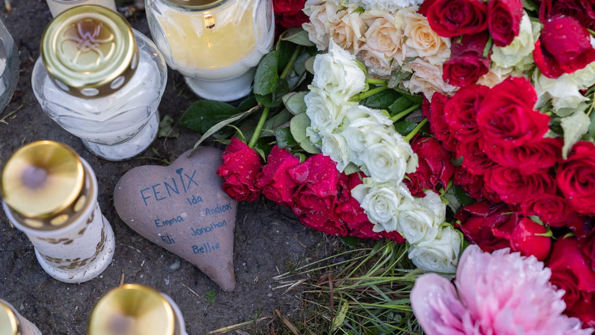 Et hjerteformet minnesmerke med navn og et stort blomsterarrangement med røde og hvite roser er plassert på bakken. Flere lys i glassbeholdere med gullfargede lokk står rundt minnesmerket og blomstene. Minnesmerket har navnet "Fenix" og flere andre navn skrevet på. (Bildebeskrivelsen er laget av en KI-tjeneste)