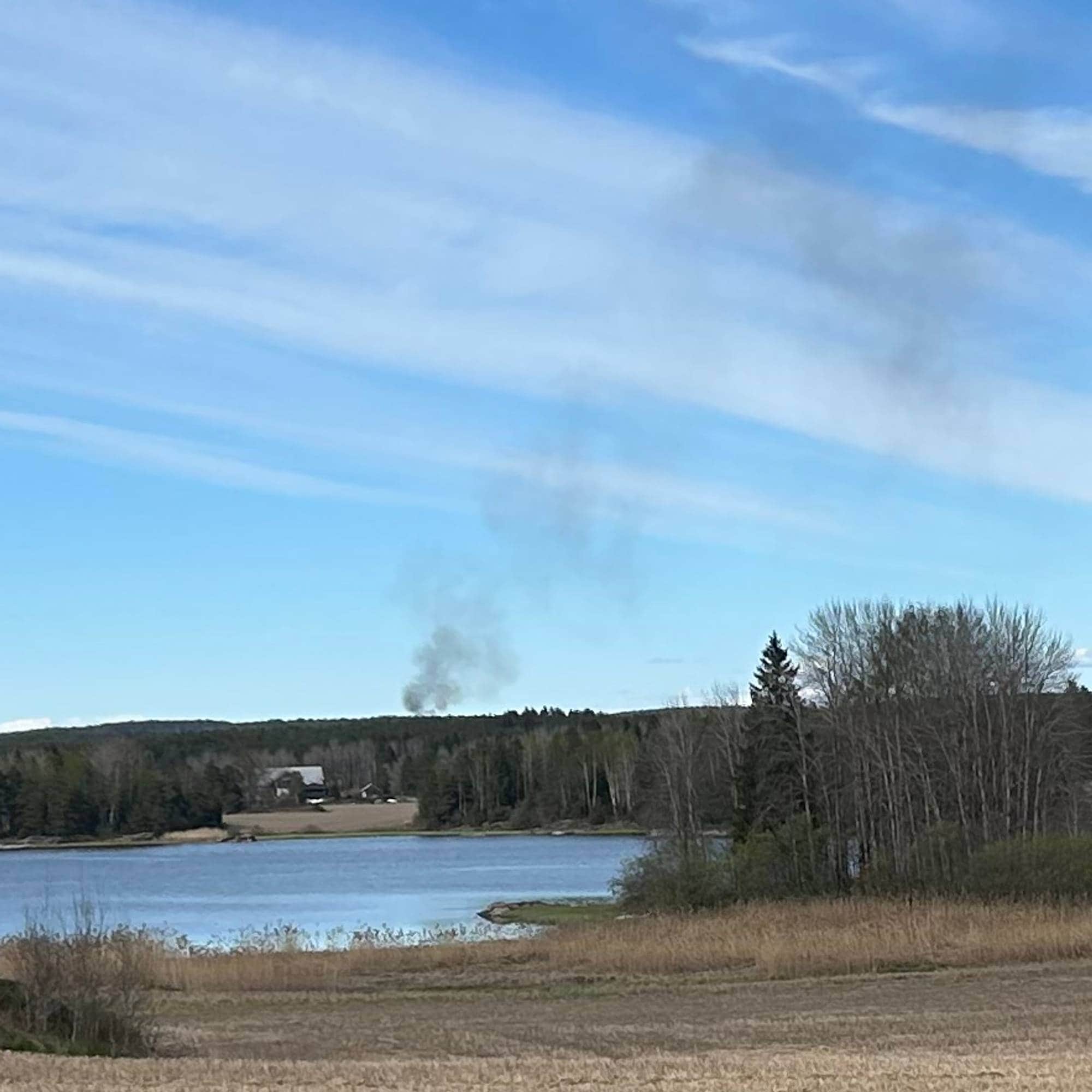 Et bilde viser et landskap med et jorde i forgrunnen, vann i midten og skog i bakgrunnen. Det er en skyfri blå himmel med tynne hvite skyer. En vei ligger langs nedre kant av bildet. (Bildebeskrivelsen er laget av en KI-tjeneste)