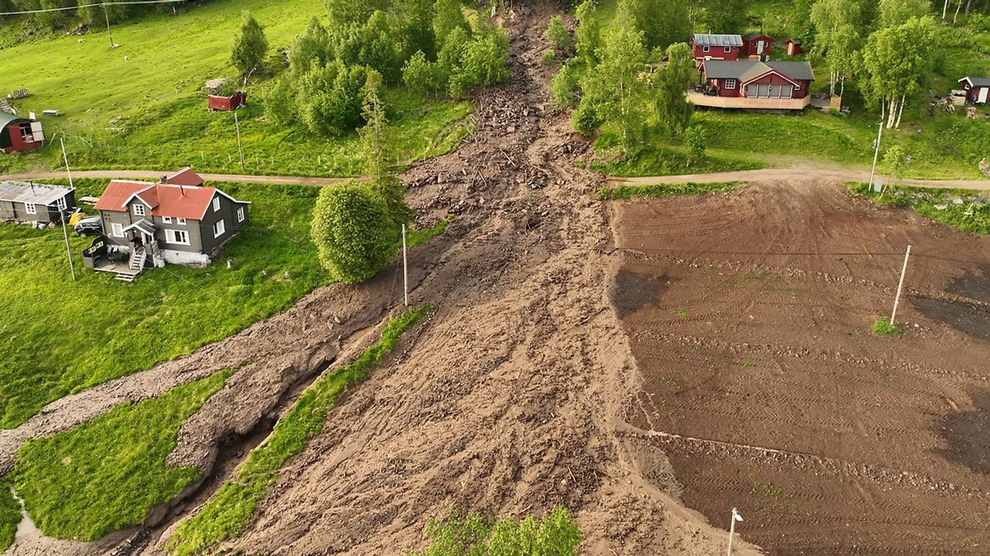 Et jordskred har gått ned en skråning og truffet flere hus. Skredet har etterlatt et bredt spor av jord og stein. Landskapet rundt er grønt med trær og gress. Det er også et dyrket jordstykke synlig. (Bildebeskrivelsen er laget av en KI-tjeneste)