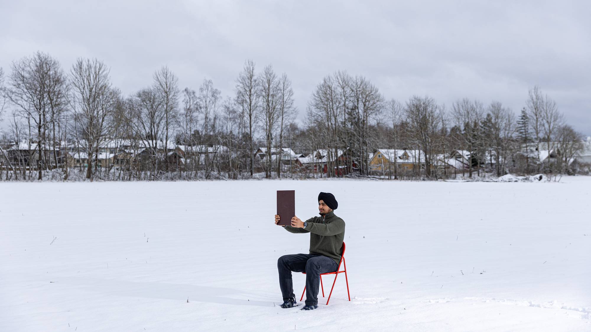 Et stort bilde av Gurveer som sitter på en rød klappstol i snøen ute på et jorde. Vi ser han holder opp speilet som han ser inn i. Han kaster en skygge i snøen, og han smiler fortsatt.