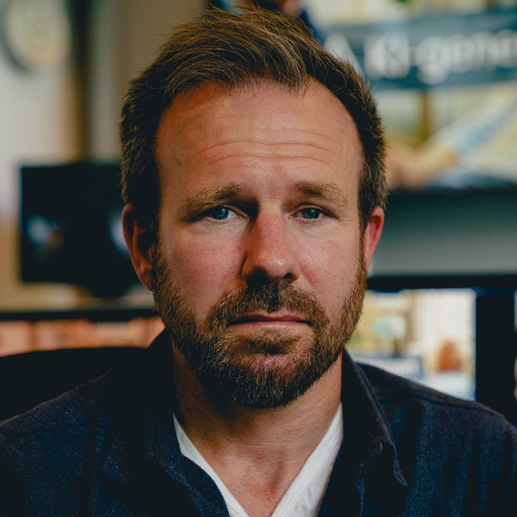 Portrett av en mann som ser i kamera. Han har brunt hår og skjegg og er klegg i en skjorte og t-skjorte