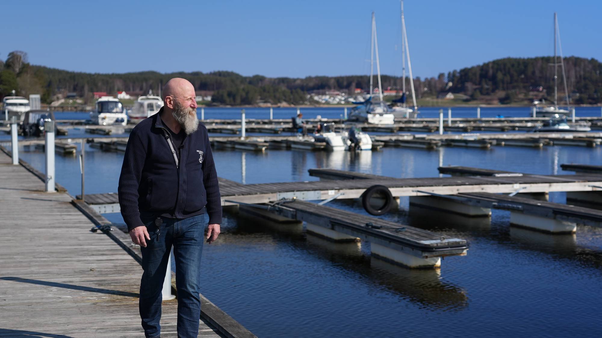 En mann går på en brygge ved en marina.
Han har på seg mørk jakke og jeans.
I bakgrunnen er det flere båter og seilbåter fortøyd.
Omgivelsene viser en klar blå himmel og et rolig vann. (Bildebeskrivelsen er laget av en KI-tjeneste)