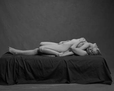 To nakne kvinner med blondt hår ligger på en mørk seng. Den ene personen ligger på den andre med ryggen til den andre og beina bøyd. Den øverste kvinnen har den ene hånda i skrittet, mens den underste kvinnen holder den ene hånda på den andres bryst. Øynene er lukket.
