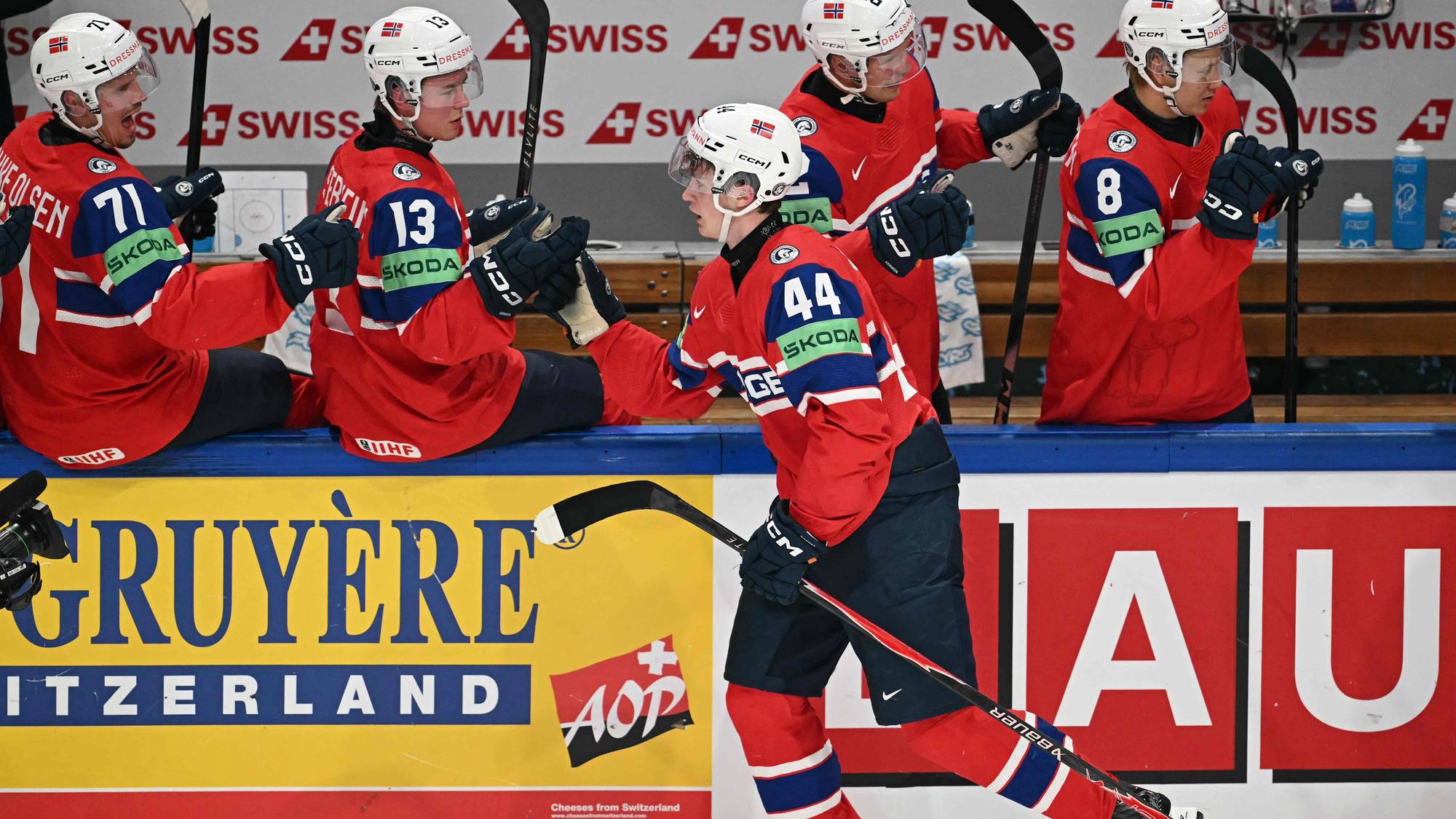 Norske ishockeyspillere i røde drakter sitter på benken under en kamp. En spiller, nummer 44, går ut på isen. Bak spillerne er det en reklamevegg med teksten "GRUYÈRE SWITZERLAND". (Bildebeskrivelsen er laget av en KI-tjeneste)
