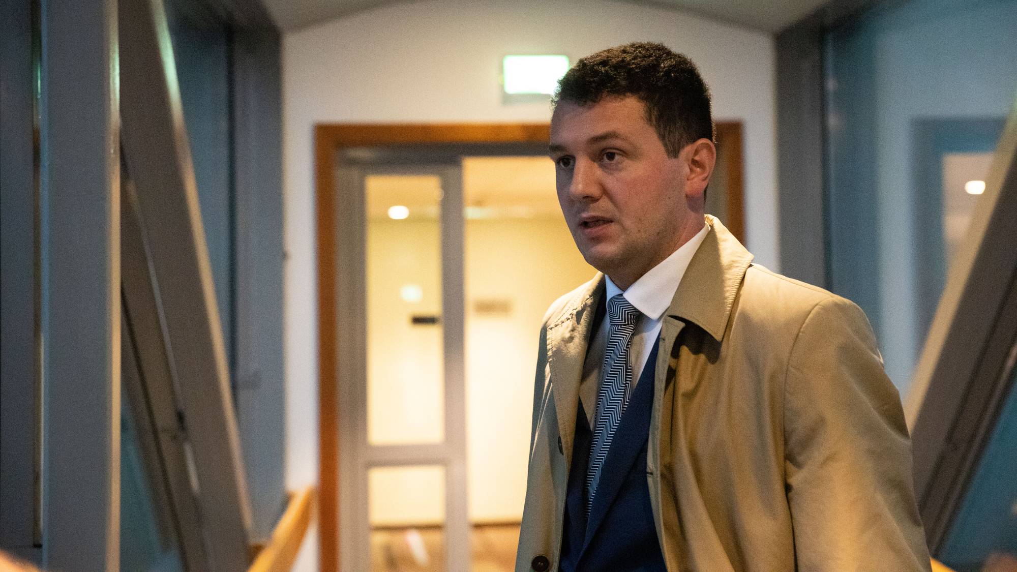 En mann i dress og beige frakk står i en korridor. (Bildebeskrivelsen er laget av en KI-tjeneste)