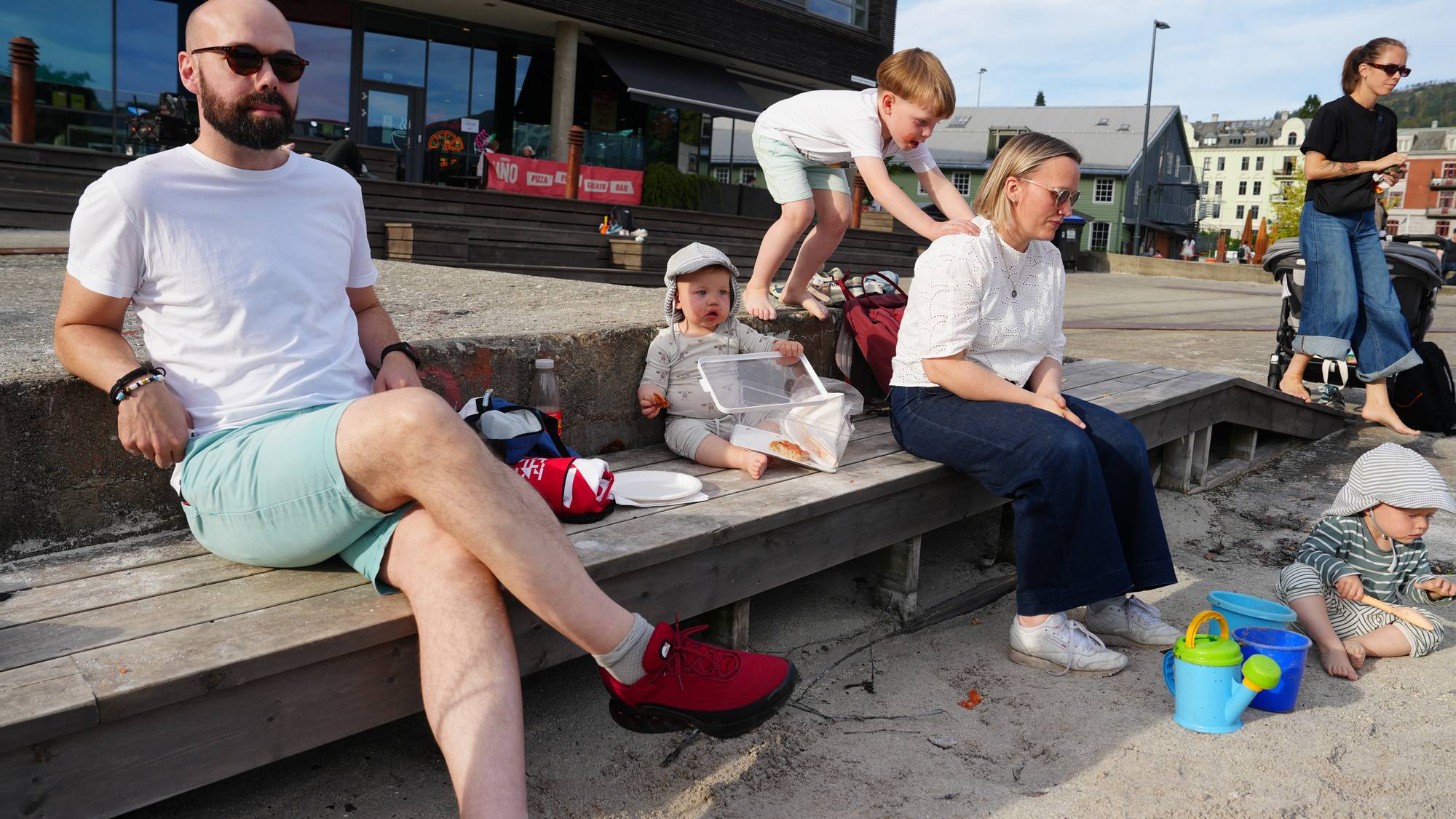 Mor, far, ein liten son og ein baby sit på ein benk. Guten står bak mora og lener seg med hendene mot ryggen hennar.