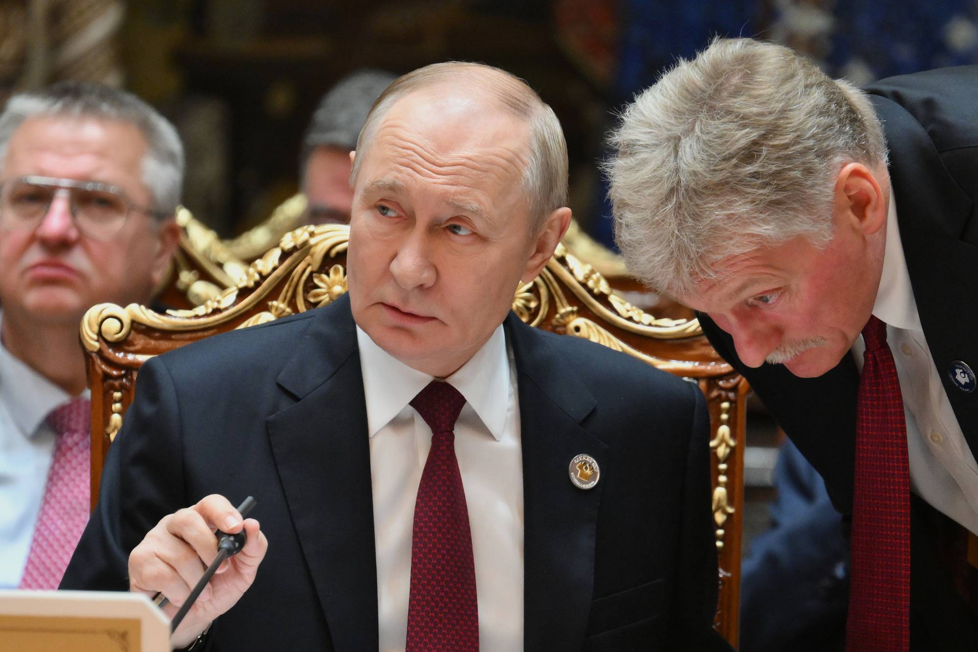 Vladimir Putin sitter ved et bord mens Dmitrij Peskov lener seg frem for å snakke med ham. (Bildebeskrivelsen er laget av en KI-tjeneste)