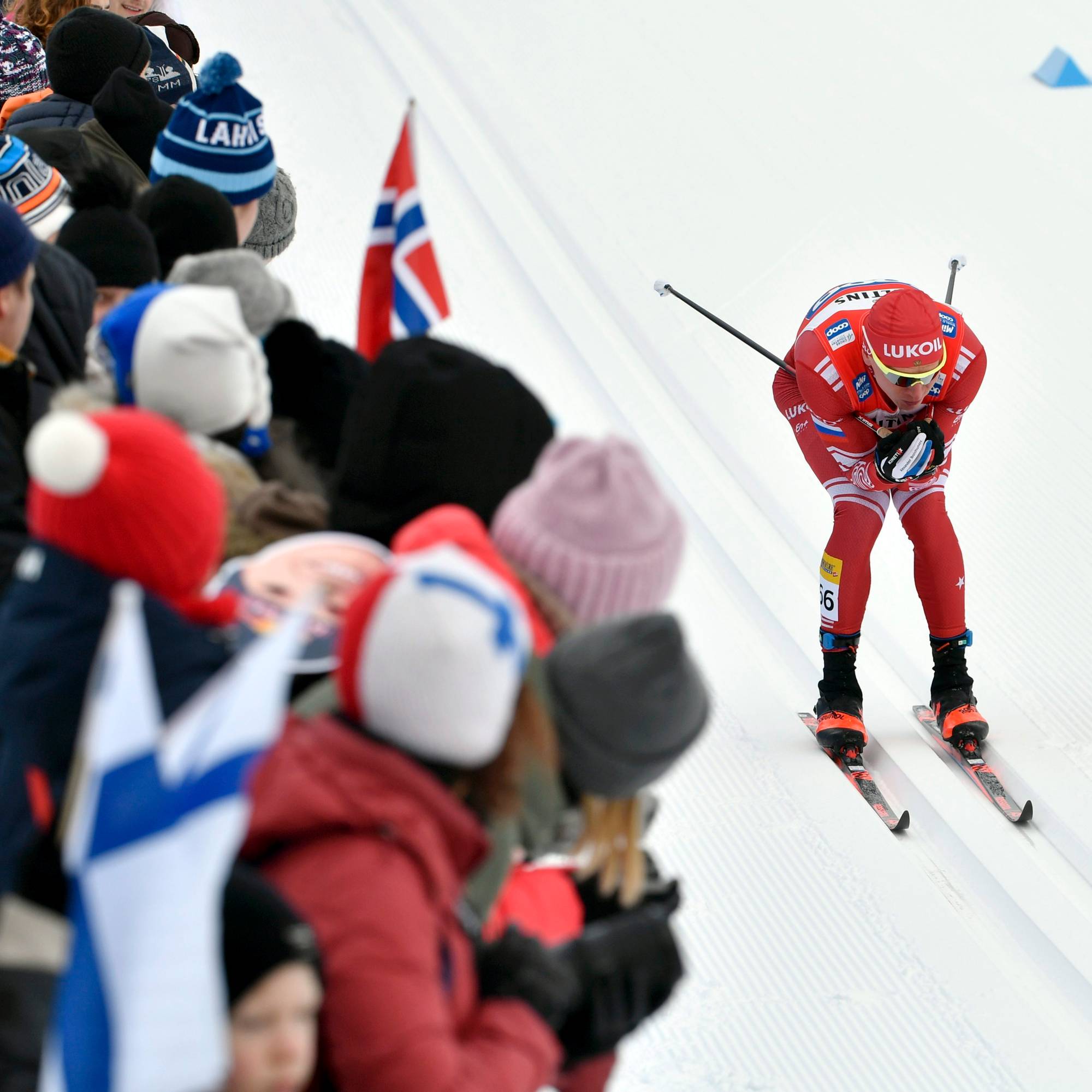 Langrennsløper i rød drakt går i løypa foran tilskuere med norske og finske flagg. (Bildebeskrivelsen er laget av en KI-tjeneste)
